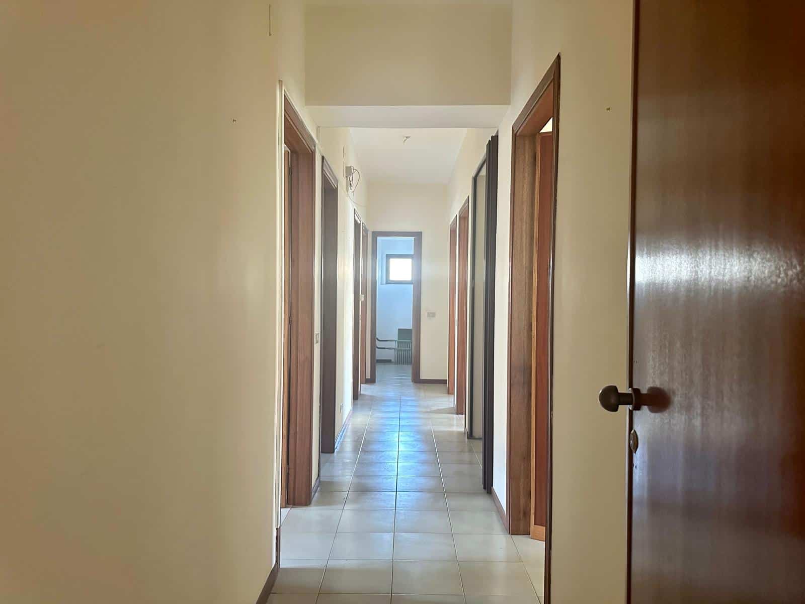 Corridor di un appartamento con pareti beige, pavimento in piastrelle e porte in legno, luminoso grazie alla finestra alla fine del corridoio, ideale per case in vendita e affitti.