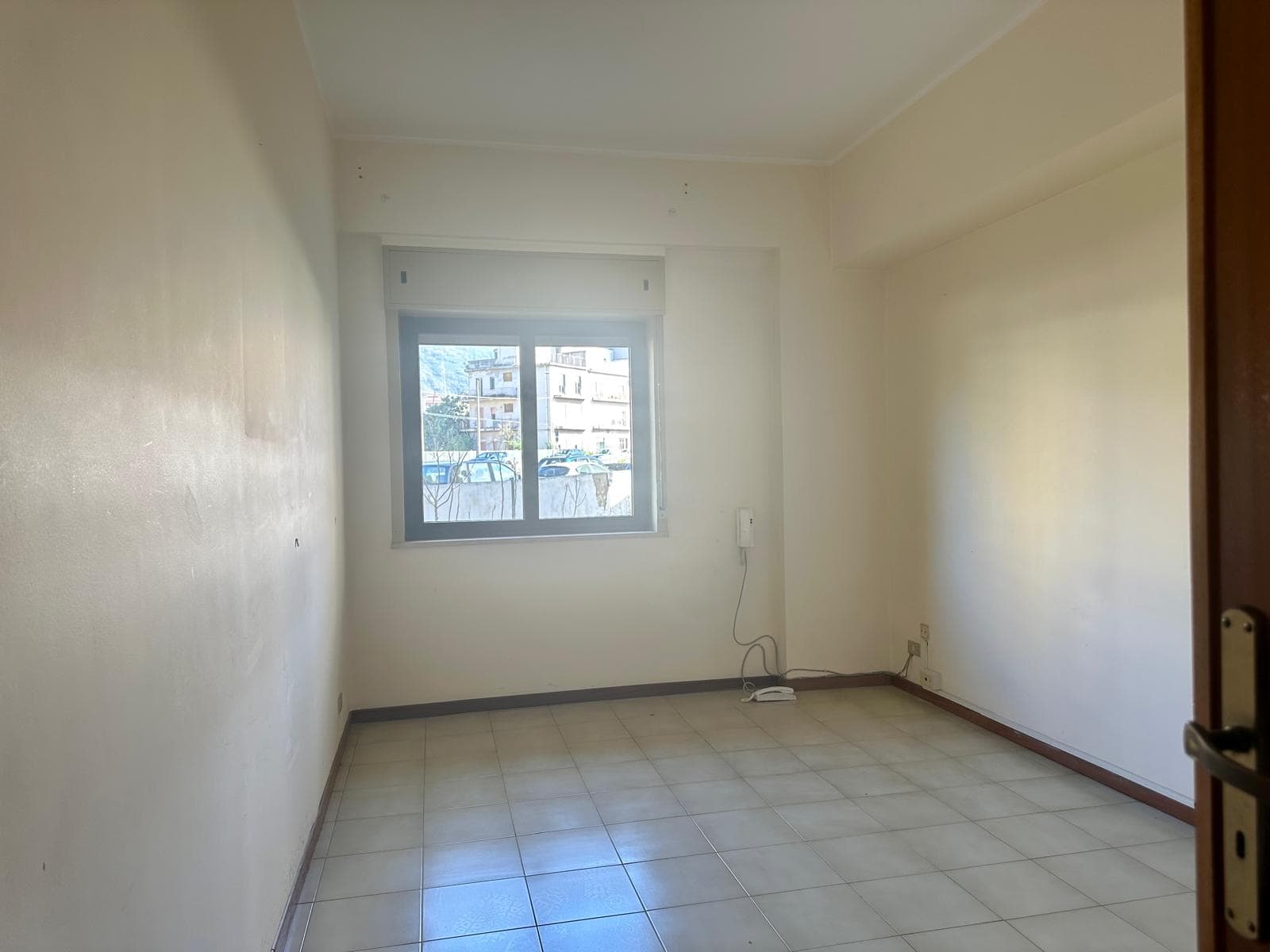 Camera vuota in appartamento in vendita, luminosa con finestra che offre vista su edifici circostanti, pavimento in piastrelle beige e pareti bianche. Ideale per ristrutturare e personalizzare.
