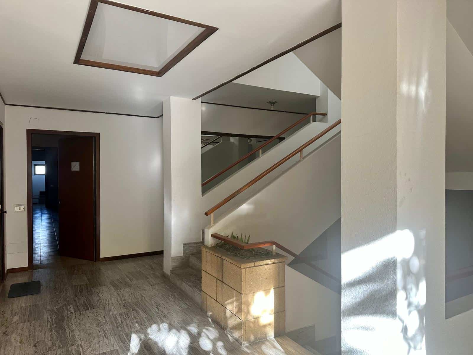 Moderno ingresso di casa con soffitto a design quadrato in cristallo, pareti bianche, pavimento in gres effetto legno, scala interna con ringhiera in legno, luce naturale che crea riflessi e ombre.