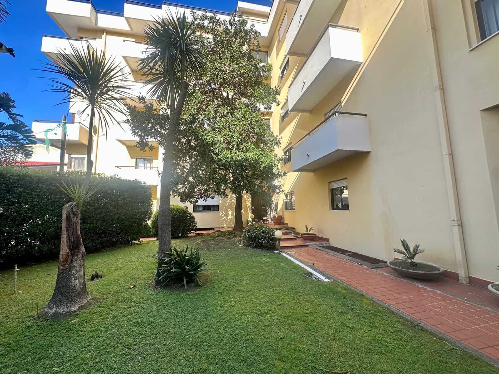 Prato curato con alberi e piante in un cortile condominiale soleggiato, edificio residenziale con balconi, ambienti esterni tranquilli e arredi estetici.