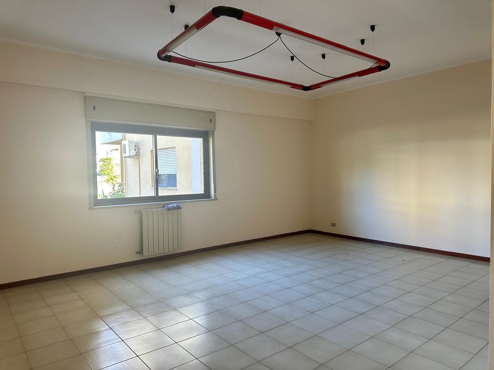 Soggiorno vuoto con pareti bianche, finestra grande che lascia entrare molta luce naturale, pavimento in piastrelle beige e un radiatore sotto la finestra, pronto per arredare.