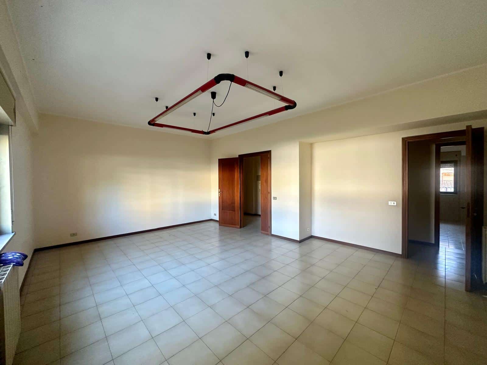 Spazio soggiorno vuoto con pareti chiare e pavimento in ceramica, grandi finestre per molta luce naturale, porte in legno e attuale installazione di luce hitop a soffitto.