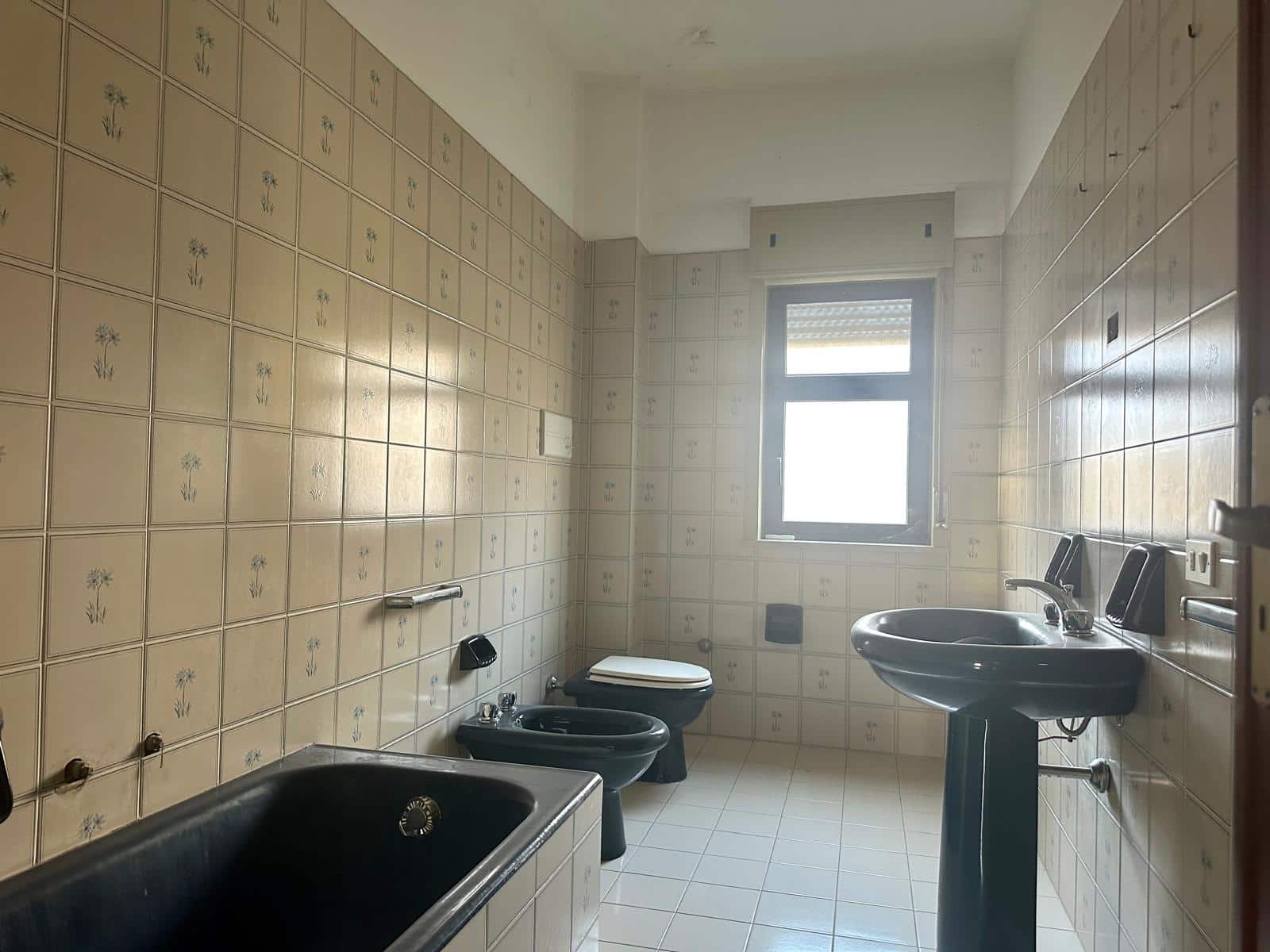 Rustico bagno con piastrelle beige con fiori blu e bianco, lavabo, bidet e WC neri, grande finestra per luce naturale, dettagli di arredamento in stile classico, ideale per casa di campagna o ristrutturazione residence.