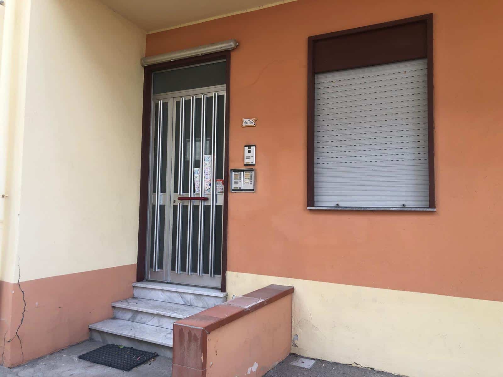 Porta d'ingresso in metallo con sbarre, con patio e finestre con tapparelle, facciata di casa con colori caldi, dettagli di sicurezza e accesso all'appartamento.