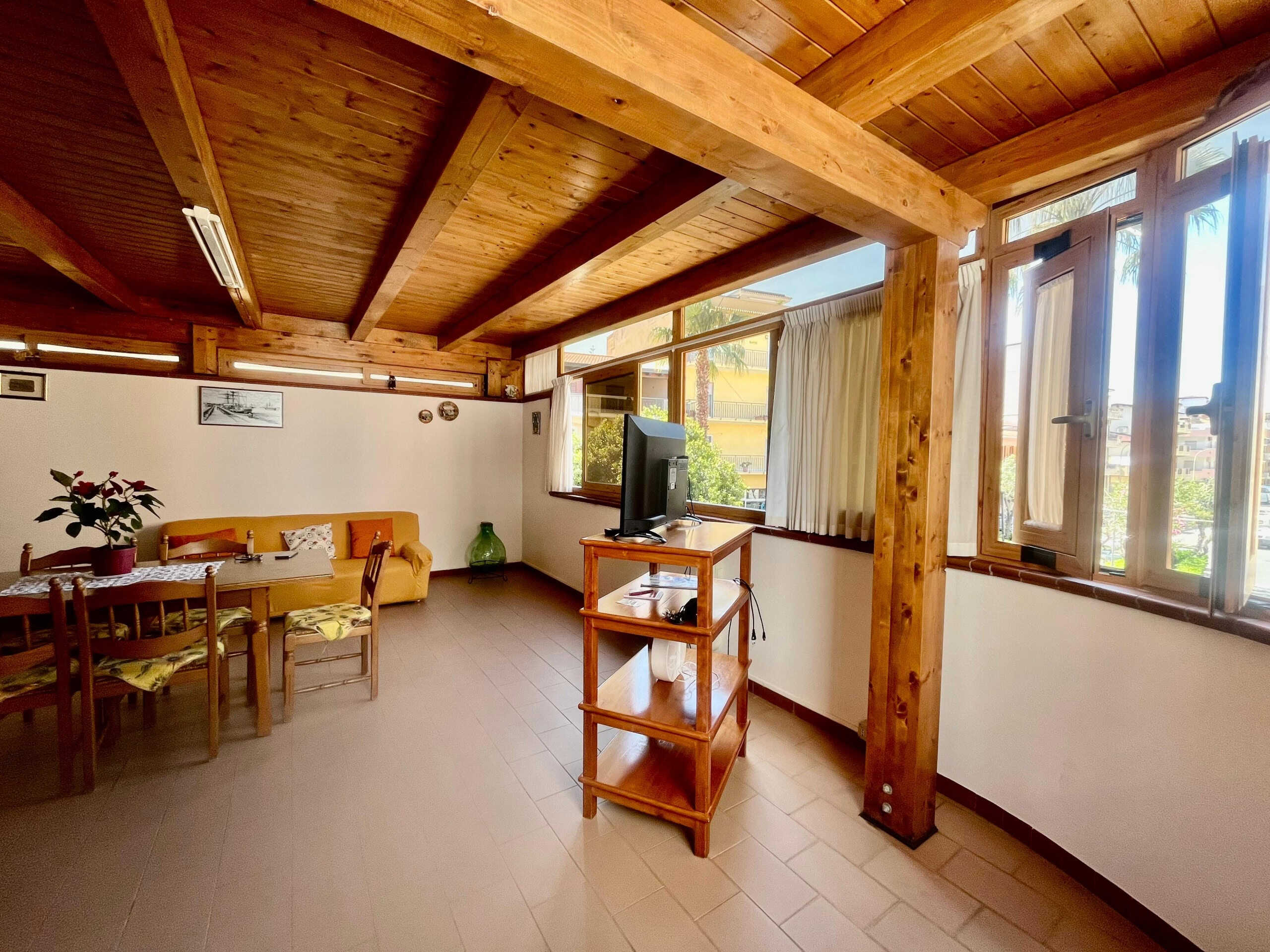 Accogliente soggiorno con pareti bianche, soffitto in legno a vista e grandi finestre che offrono luce naturale, arredato con divano, tavolo e TV, ideale per relax e convivialità.