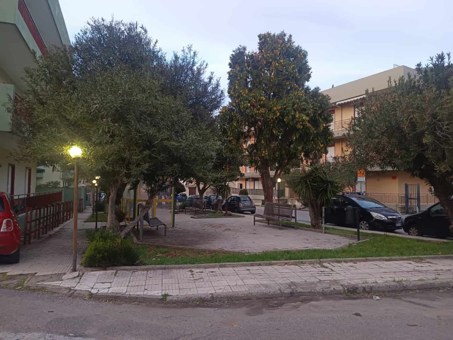 Ampia area parcheggio esterna circondata da alberi e panchine, in zona residenziale tranquilla con edifici multifamiliari. Luogo ideale per residenti e visitatori.