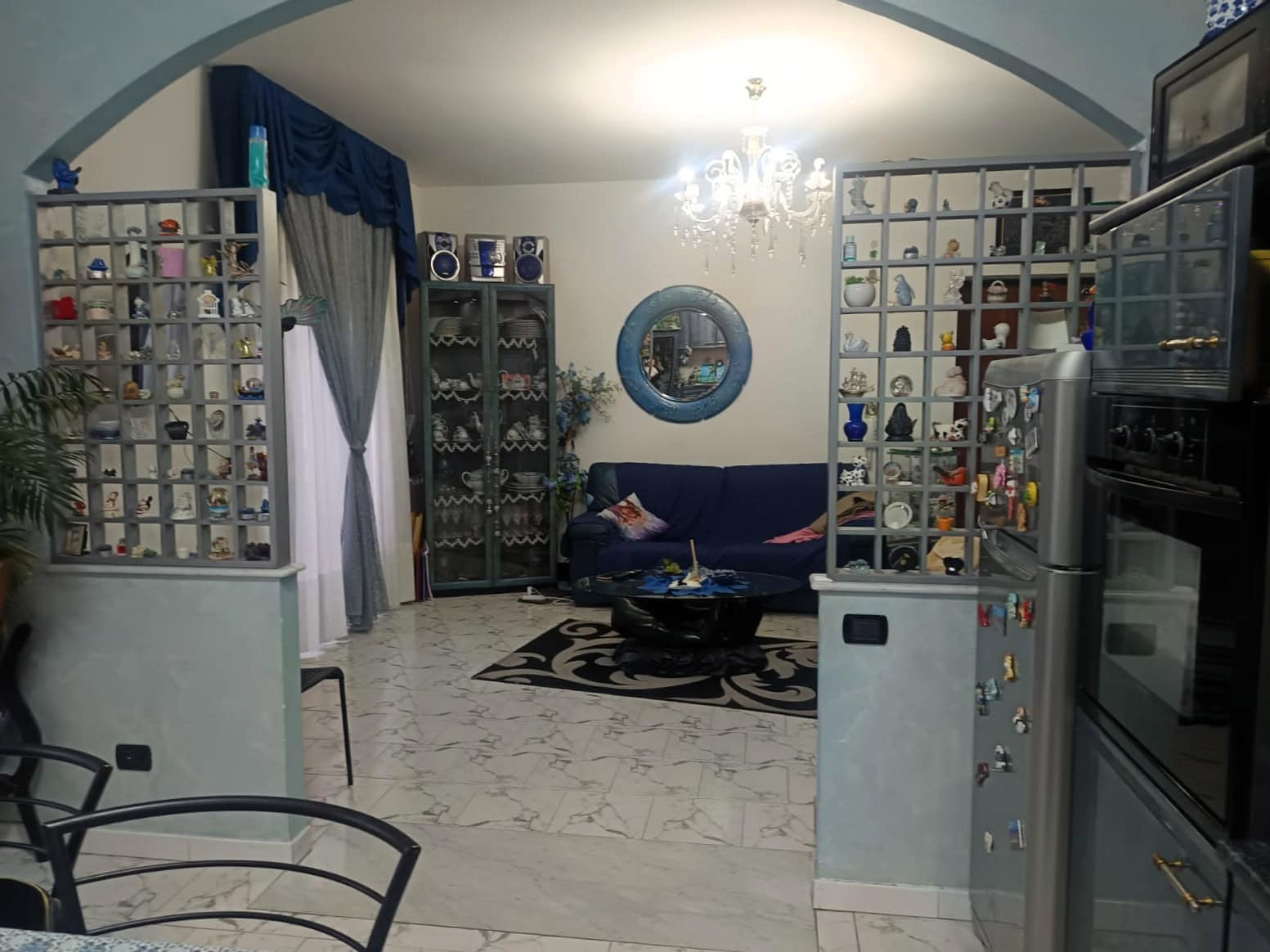 1. Divano blu in soggiorno con arredamento elegante e scaffalature con oggetti decorativi, illuminata da lampadario centrale.