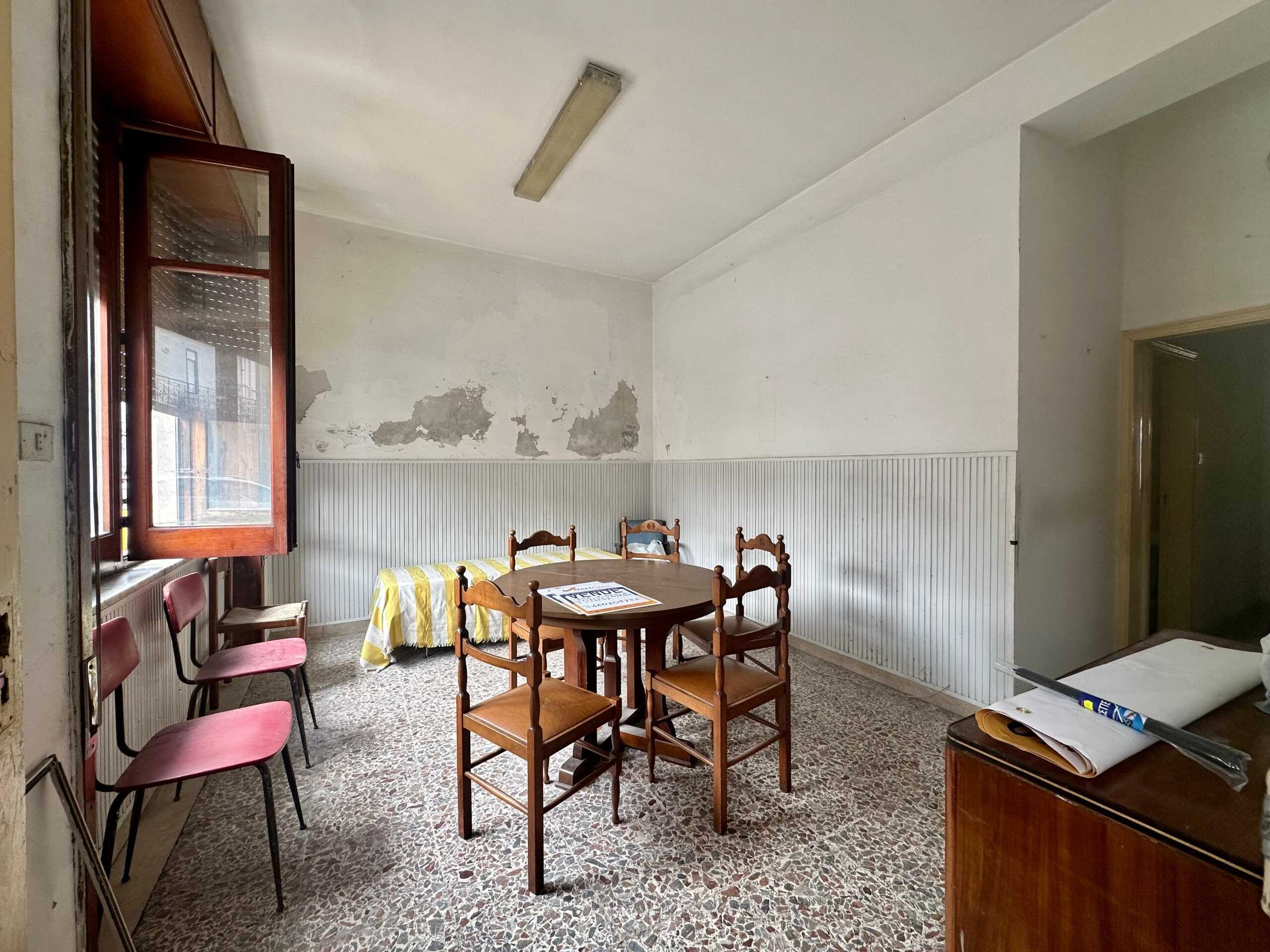 Stanza di abitazione con pareti usurate e mobili d'epoca, ambiente da ristrutturare, luce naturale attraverso finestra aperta, pavimento in terrazzo, stile rustico e vintage, potenziale per rinnovamento.