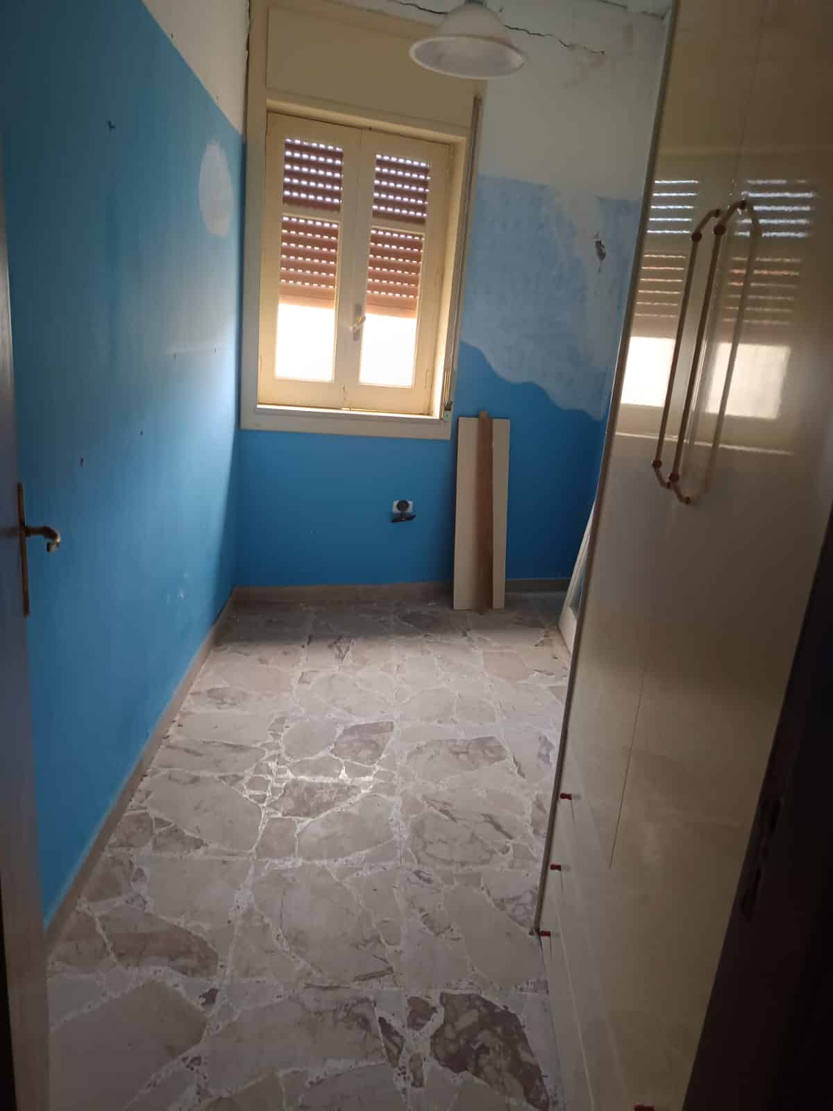 Antico bagno in ristrutturazione con pareti blu e finestra, pavimento in marmo e armadio a muro bianco, pronta per essere arredato.