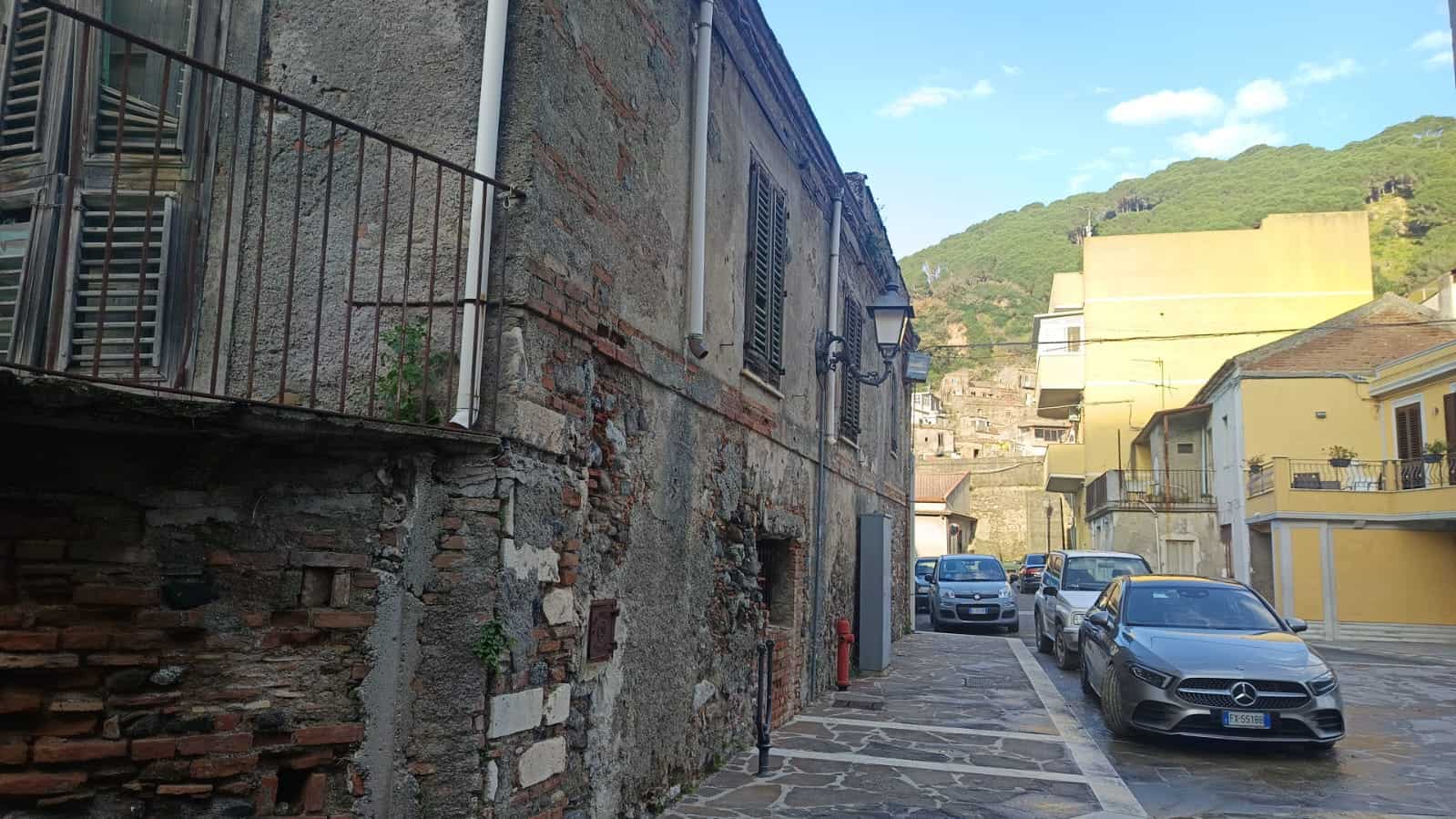 Vecchio edificio in pietra con finestre con persiane, vicolo cittadino con auto moderne, contesto urbano vicino a colline verdi e cielo sereno - NextCasa.