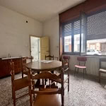Vecchia casa con soggiorno spazioso, tavolo in legno, sedie in stile vintage e grandi finestre con vista sulle strade circostanti, caratterizzata da un ambiente accogliente e un tocco rustico.