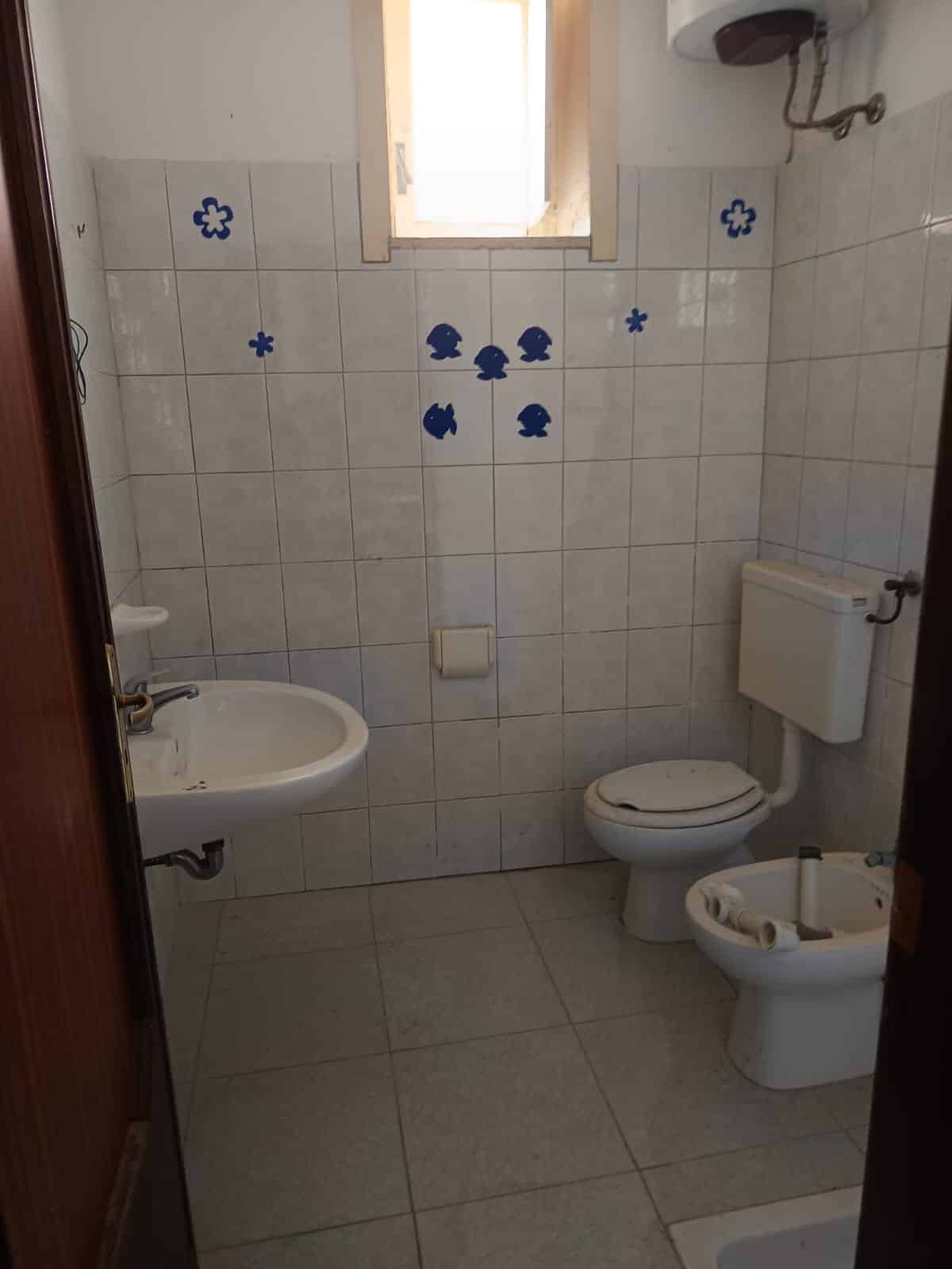 Lavabo e water nel bagno di un'abitazione, pareti decorate con motivi di pesci e fiori blu, finestra naturale, piastrelle chiare, ambienti funzionali, soluzione per spazi utili e decorativi, ideale per case di vacanza o abitazioni private.