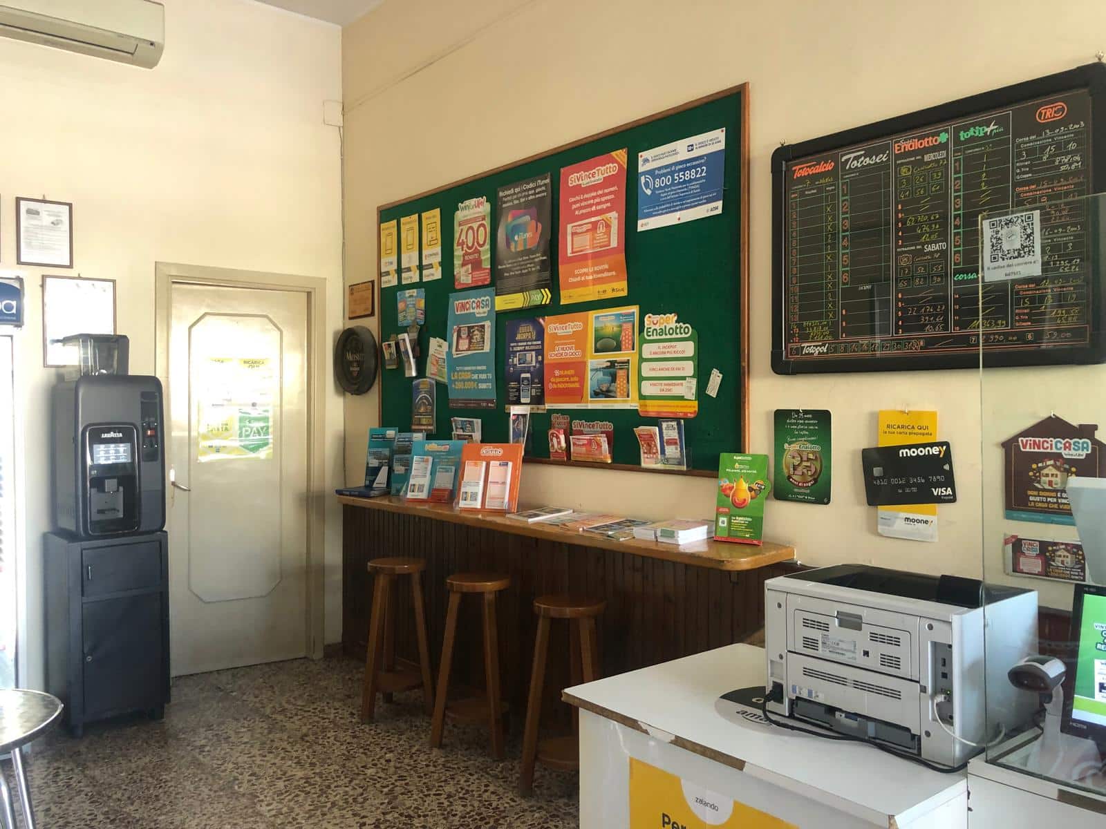 Alto testo: Punto di vendita di servizi finanziari e biglietteria con bacheca di annunci, schede di scommesse sportive, distributore di biglietti e area accoglienza clienti.