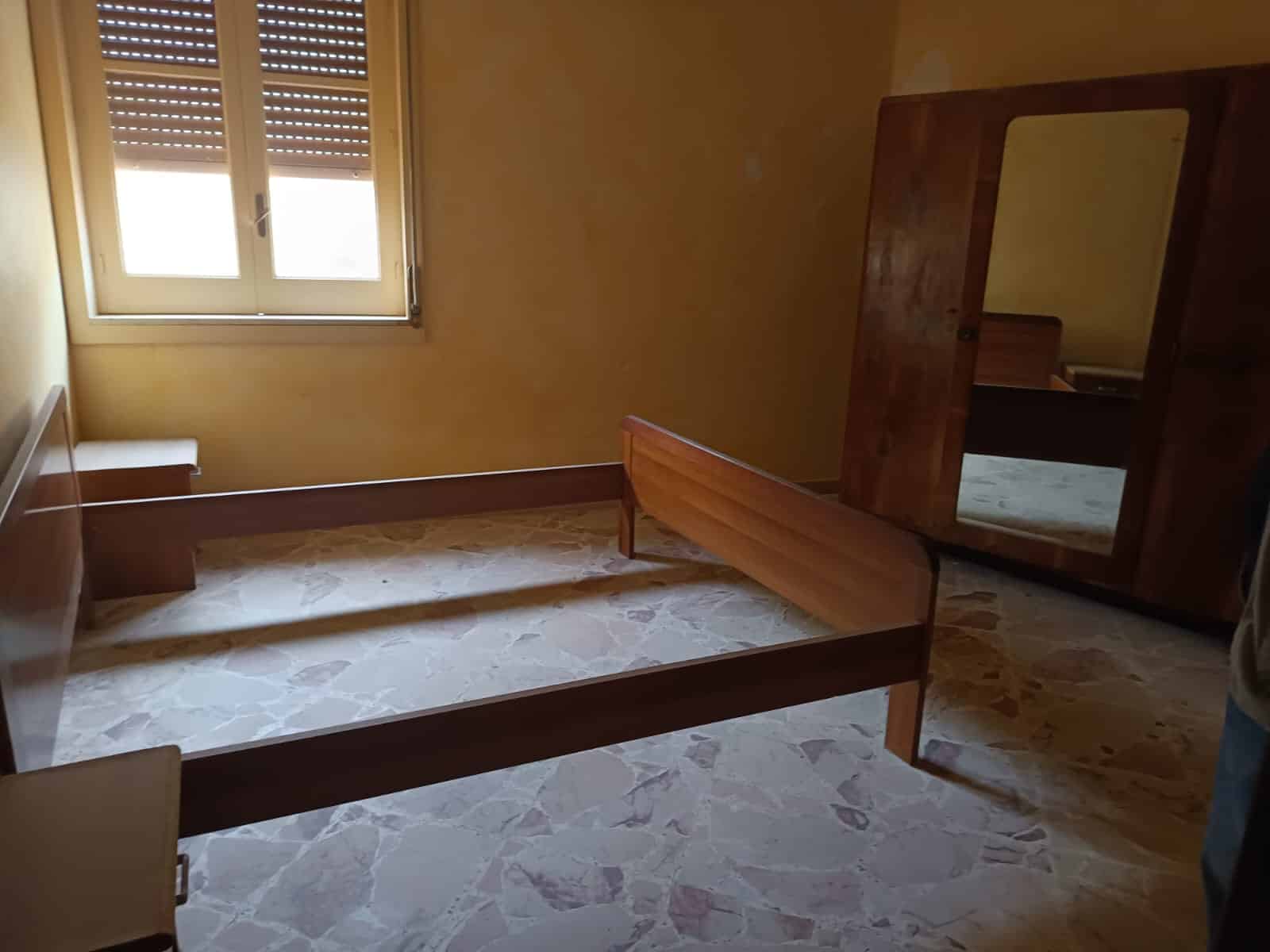 Letto in una stanza vuota con pavimento in marmo, finestra con tapparelle e armadio in legno, ambiente luminoso e semplice in stile classico.