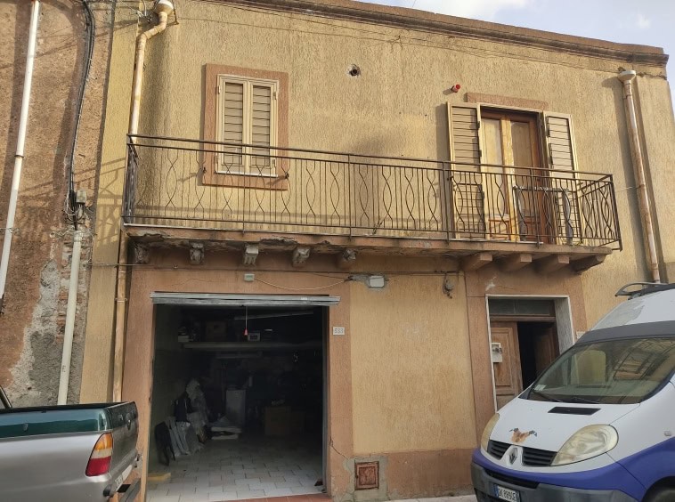 Vecchia casa in stile rustico con balcone in ferro battuto, finestre con tapparelle, ingresso con portone in legno, parcheggio e garage, pareti esterne in pietra e intonaco giallo, in zona residenziale.