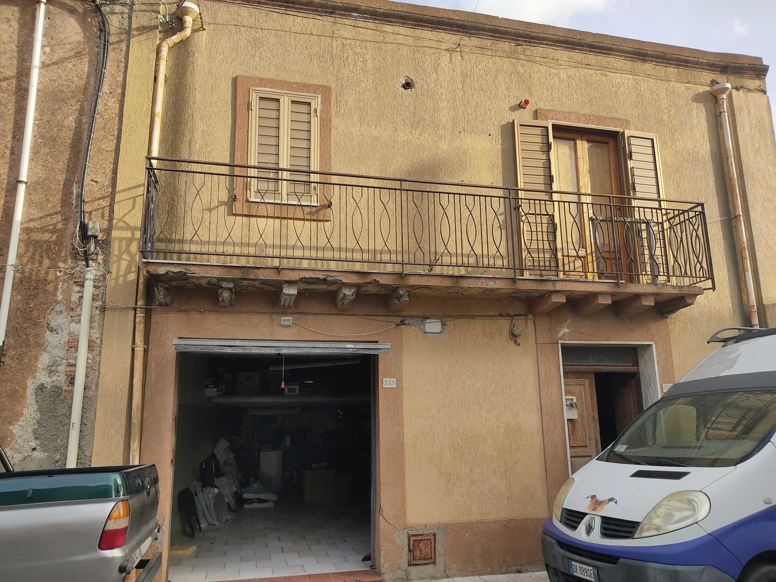 Vecchia casa in stile rustico con balcone in ferro battuto, finestre con tapparelle, ingresso con portone in legno, parcheggio e garage, pareti esterne in pietra e intonaco giallo, in zona residenziale.