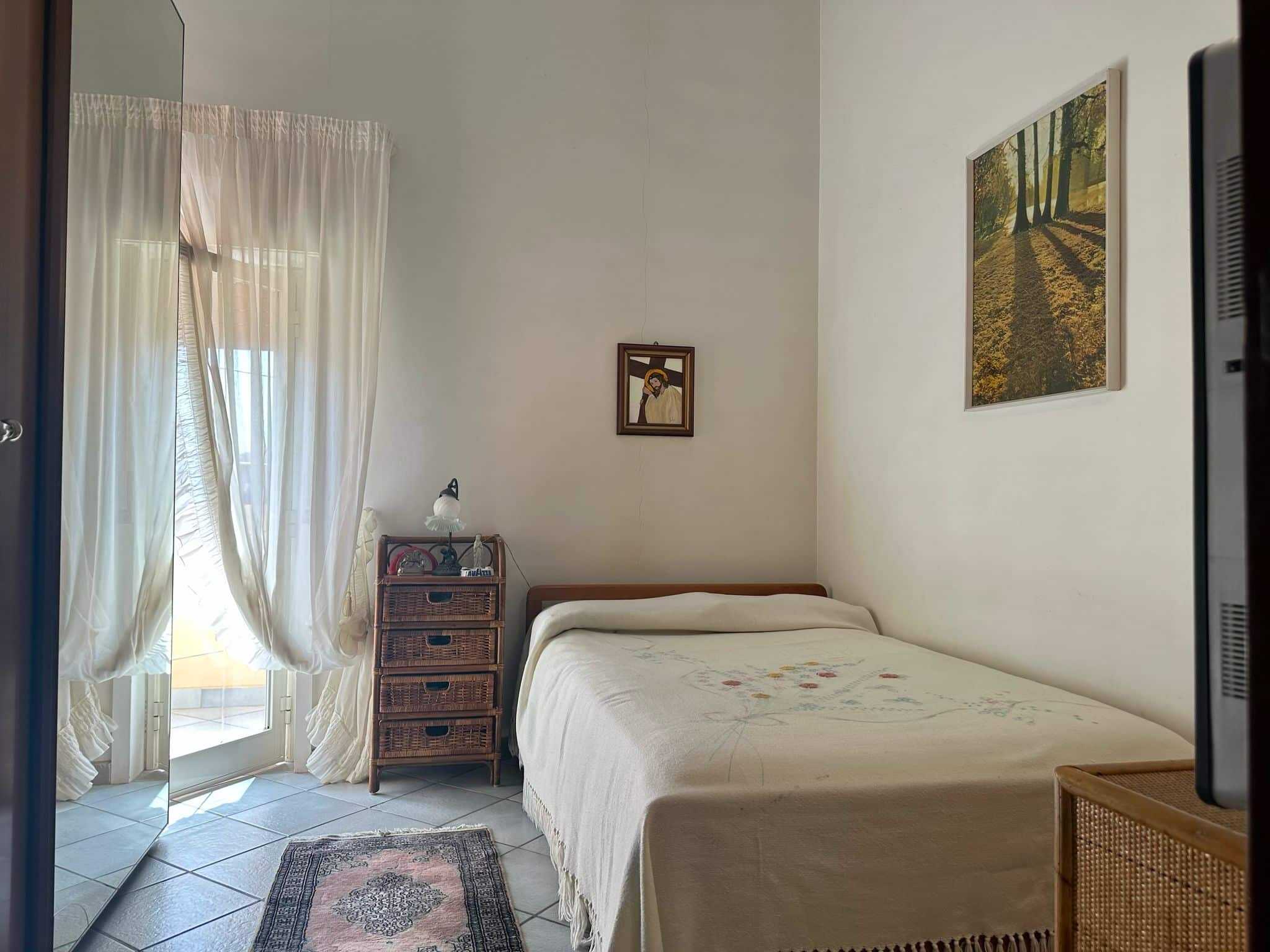 Letto matrimoniale in una stanza luminosa con pareti bianche e decorazioni floreali sulla coperta, finestra con tende leggere e quadri alle pareti, mobili in stile classico, atmosfera accogliente e tranquilla.