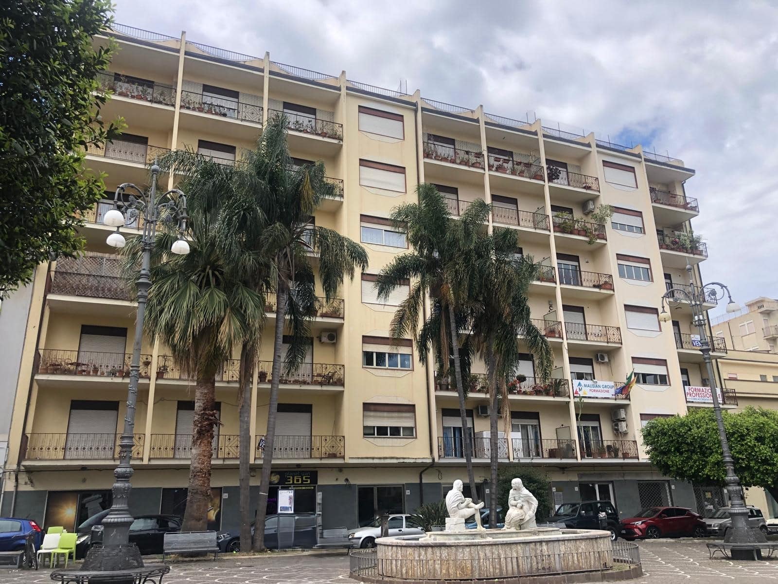 Immagine di un edificio residenziale moderno con balconi e piante, in una zona urbana con alberi e lampioni, rappresentativo di immobili di lusso e appartamenti in vendita o affitto a NextCasa.