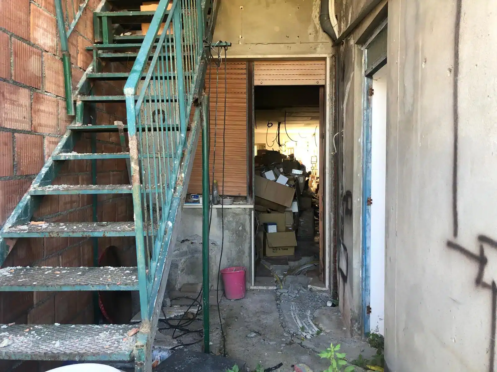 Scala esterna in metallo verde e ingresso di un'abitazione con porta aperta, visibile un interno disordinato con scatole e oggetti vari.