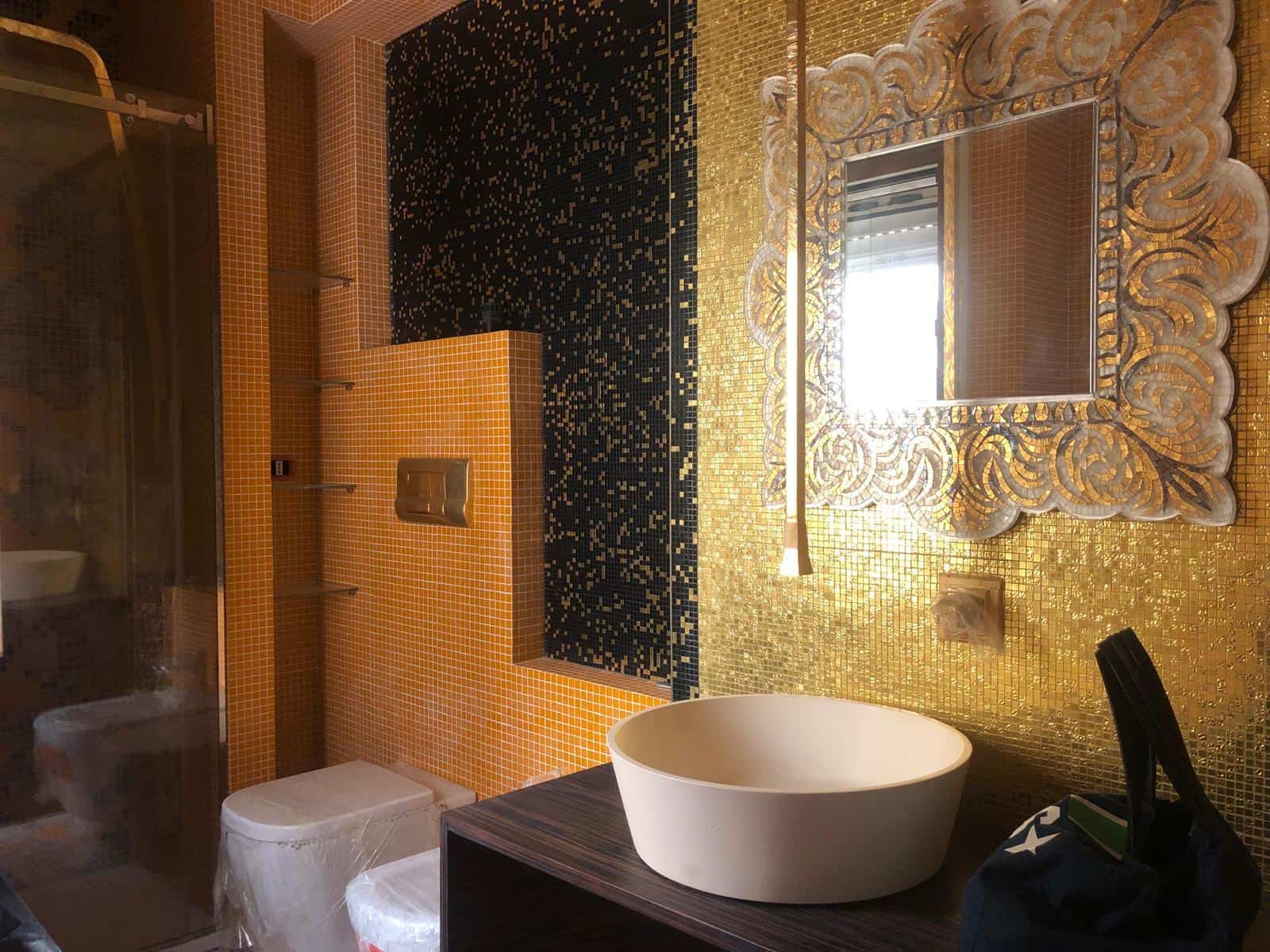 Lucido bagno di lusso con mosaico oro e dettagli in nero, lavabo rotondo in ceramica, grande specchio decorativo con cornice dorata, ambiente elegante e raffinato.
