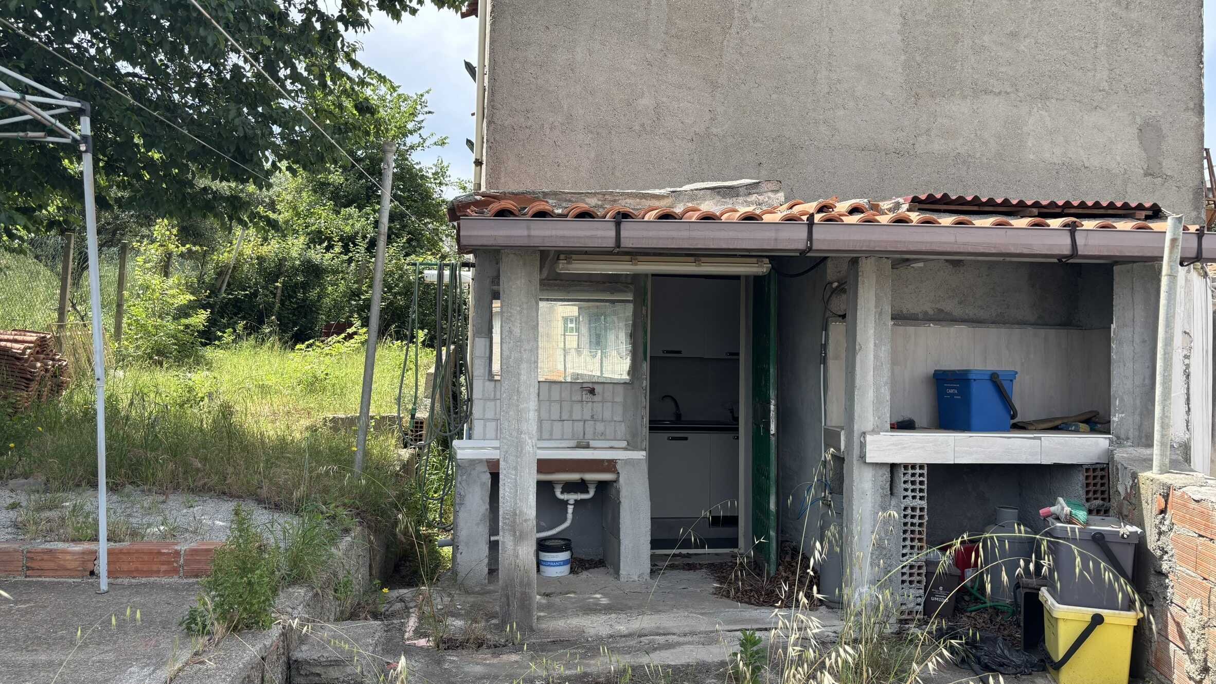 Vecchio forno esterno in costruzione, area di giardino con erba alta e piante, struttura in cemento, campagna rustica, lavoro di ristrutturazione case, spazio esterno in giardino, progetti di miglioramento abitativo, casa in fase di completamento, giardino con attrezzi e materiali da costruzione, atmosfera di rinnovamento casa.