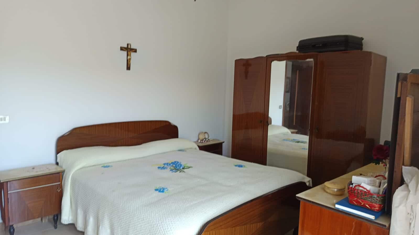 Letto matrimoniale in camera da letto con mobili in legno e arredamento classico, decorata con croce sulla parete. Ideale per case in affitto o vendita, con arredi funzionali e stile tradizionale.
