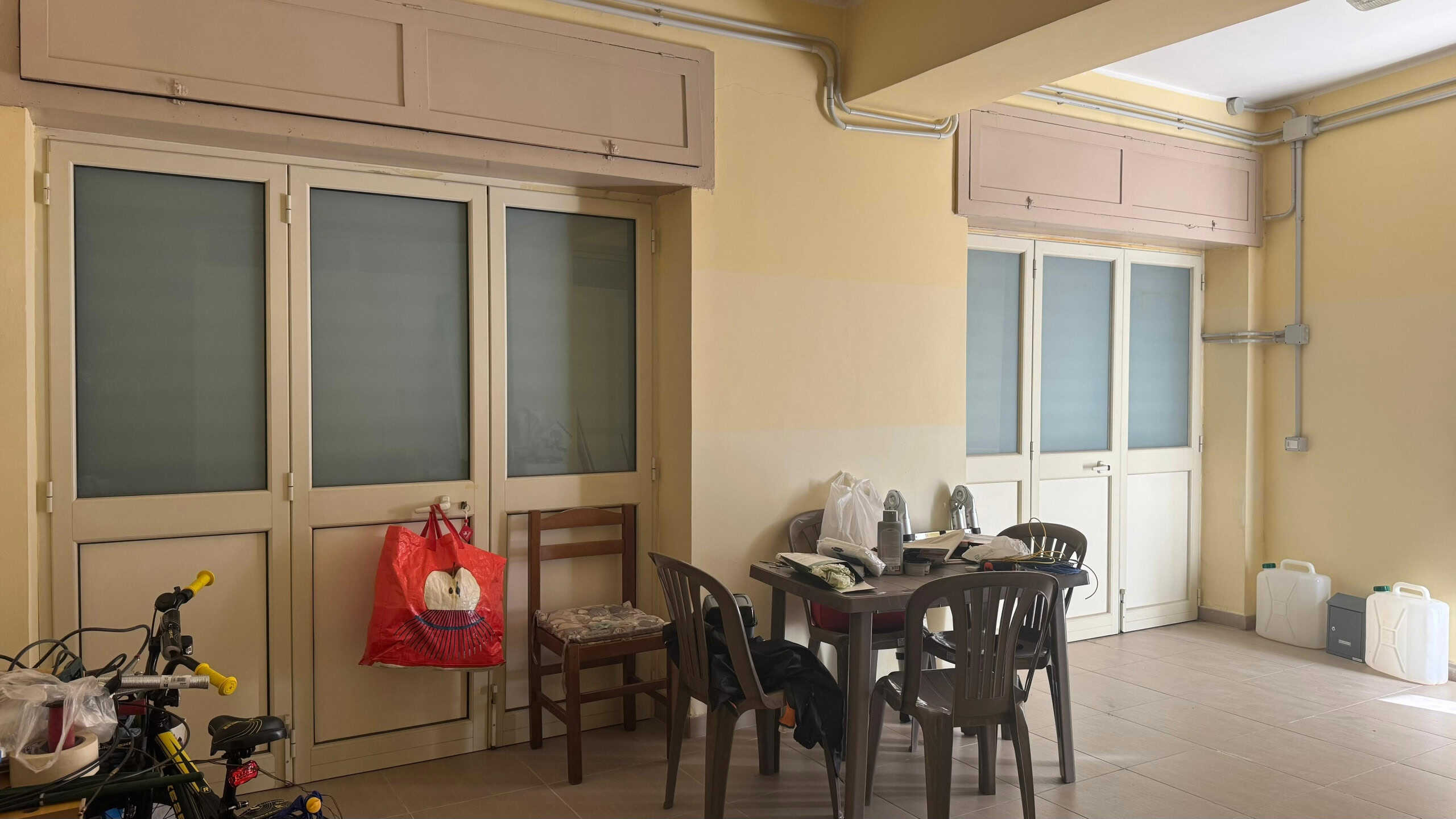 Deposito condominiale con finestra e porta scorrevole, arredato con bici, sedie e contenitori di plastica, pareti color crema, illuminazione naturale, spazio funzionale per stoccaggio e attività domestiche.