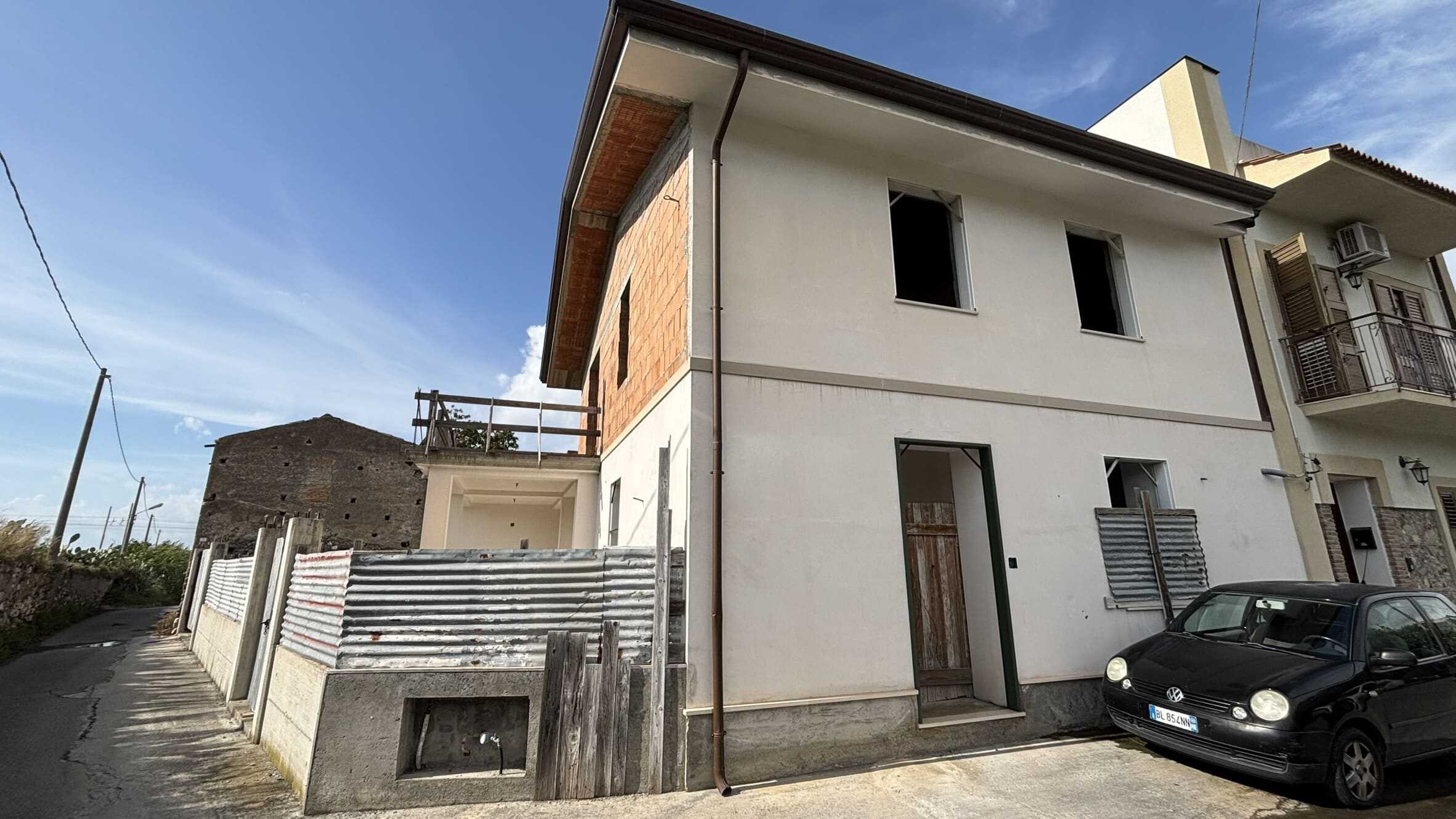 Villa in costruzione in zona residenziale, facciata esterna con pareti bianche e alcune parti in rosso mattoni, auto Volkswagen nera parcheggiata di fronte, cielo azzurro con nuvole leggere.