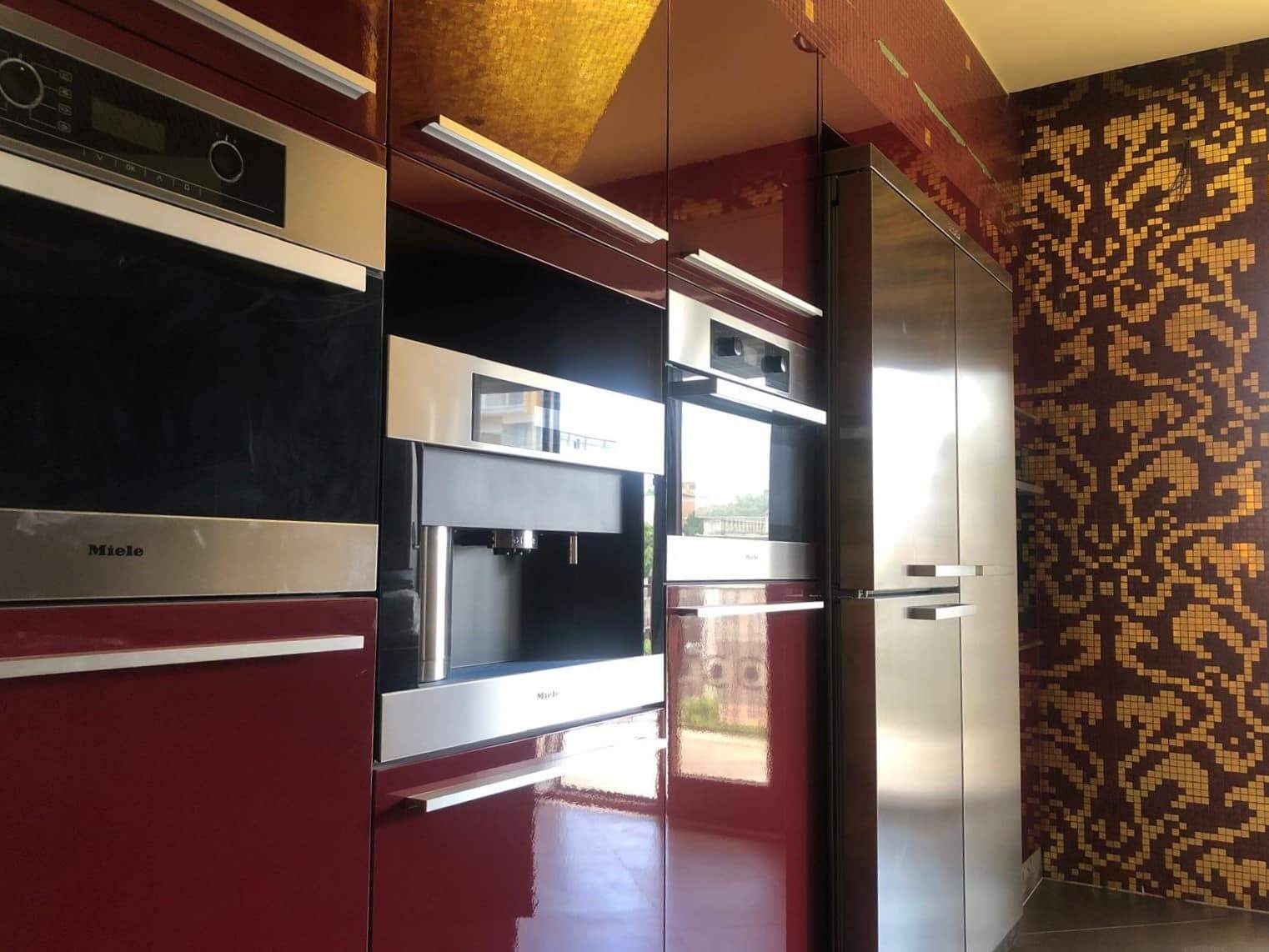 Cucina moderna con mobili in rosso lucido, elettrodomestici in acciaio inox e pareti con mosaico decorativo in toni caldi, ambiente elegante e funzionale.