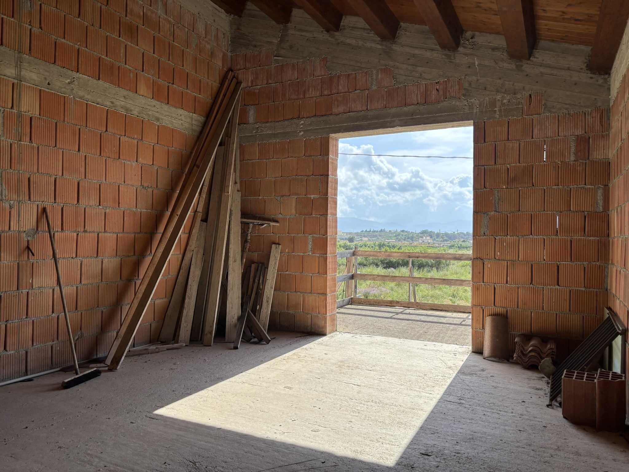 Ampio spazio in casa in costruzione con pareti in mattoni rossi e vista sul paesaggio naturale circostante. Interni ancora in fase di sviluppo, materiali da costruzione e dettagli architettonici visibili.