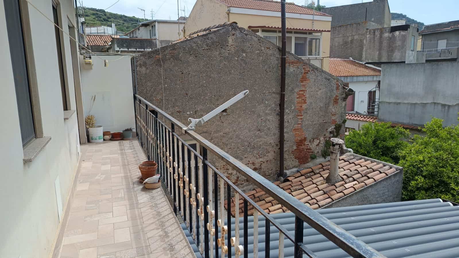 Balcone con vista su tetti e case vicine, pavimentato in piastrelle beige, con piante in vaso e segmento di ringhiera in ferro.