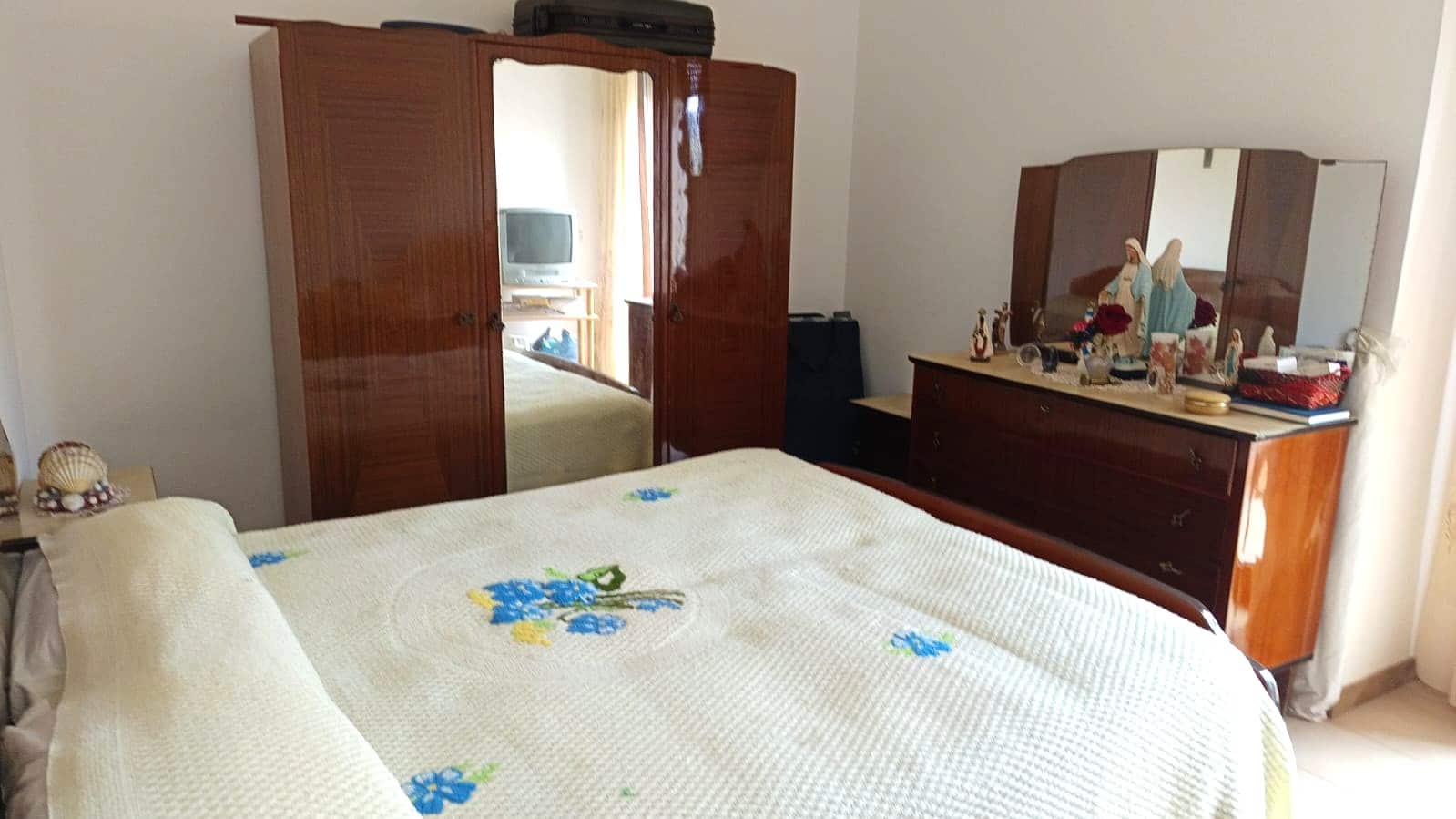 Armadio in legno con specchio, camera da letto con mobili in stile classico, arredamento elegante e decorazioni religiose e floreali, ambiente spazioso e luminoso.