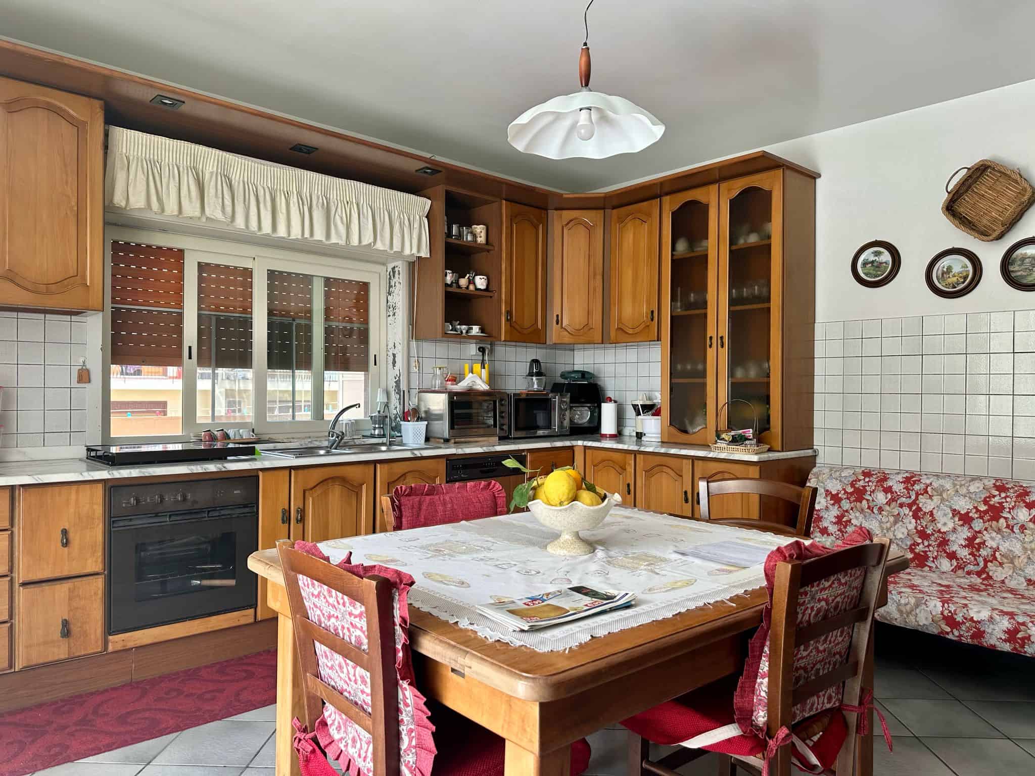 Cucina in stile classico con mobili in legno, tavolo da pranzo con tovaglia e sedie con coperture rosse, dotata di elettrodomestici e decorazioni tradizionali.