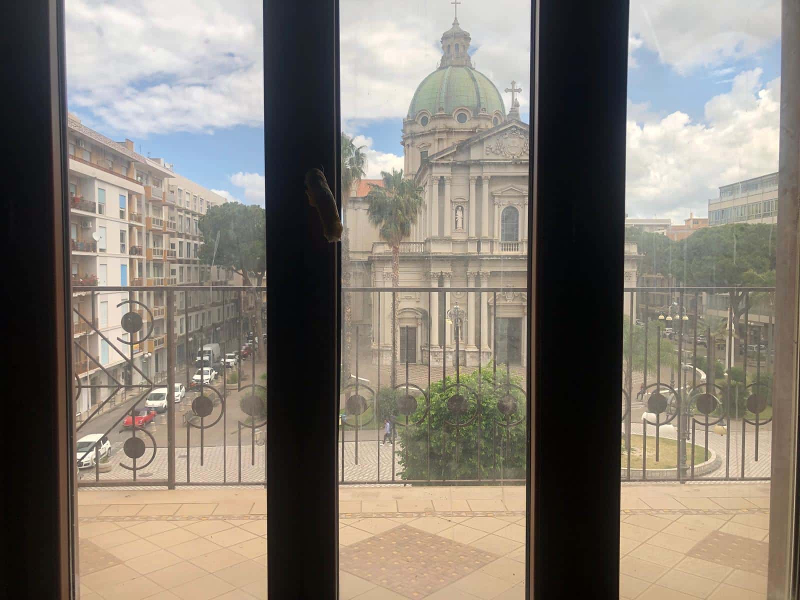 Vista panoramica dal balcone di un appartamento con l'architettura storica di una chiesa barocca al centro, fronte strada con edifici moderni, atmosfera luminosa e accogliente, ideale per affitti o residenza.