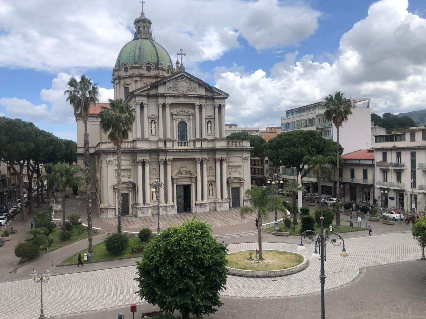 Elegante chiesa barocca con cupola verde e facciata decorata, situata in un piazzale con alberi, lampioni e persone. Panoramica della zona urbana con edifici moderni e vegetazione, cielo nuvoloso.
