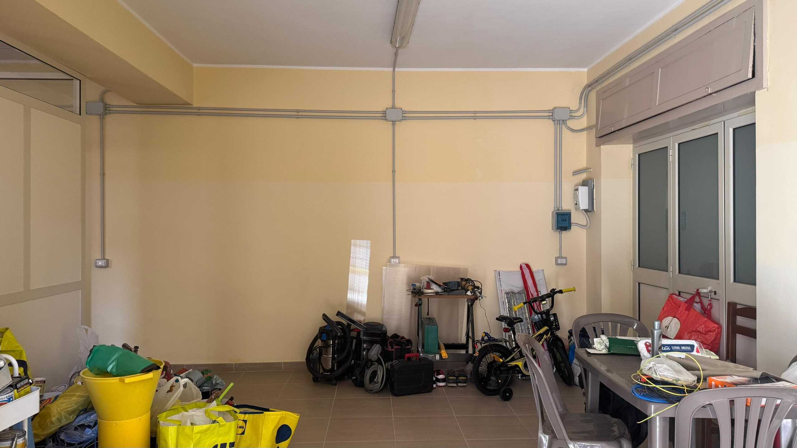 Finestra sulla parete con impianto elettrico visibile, arredamento di una stanza con giocattoli, attrezzi e materiali per lavori domestici, ambiente funzionale e pratico, ideale per abitazioni e ristrutturazioni.