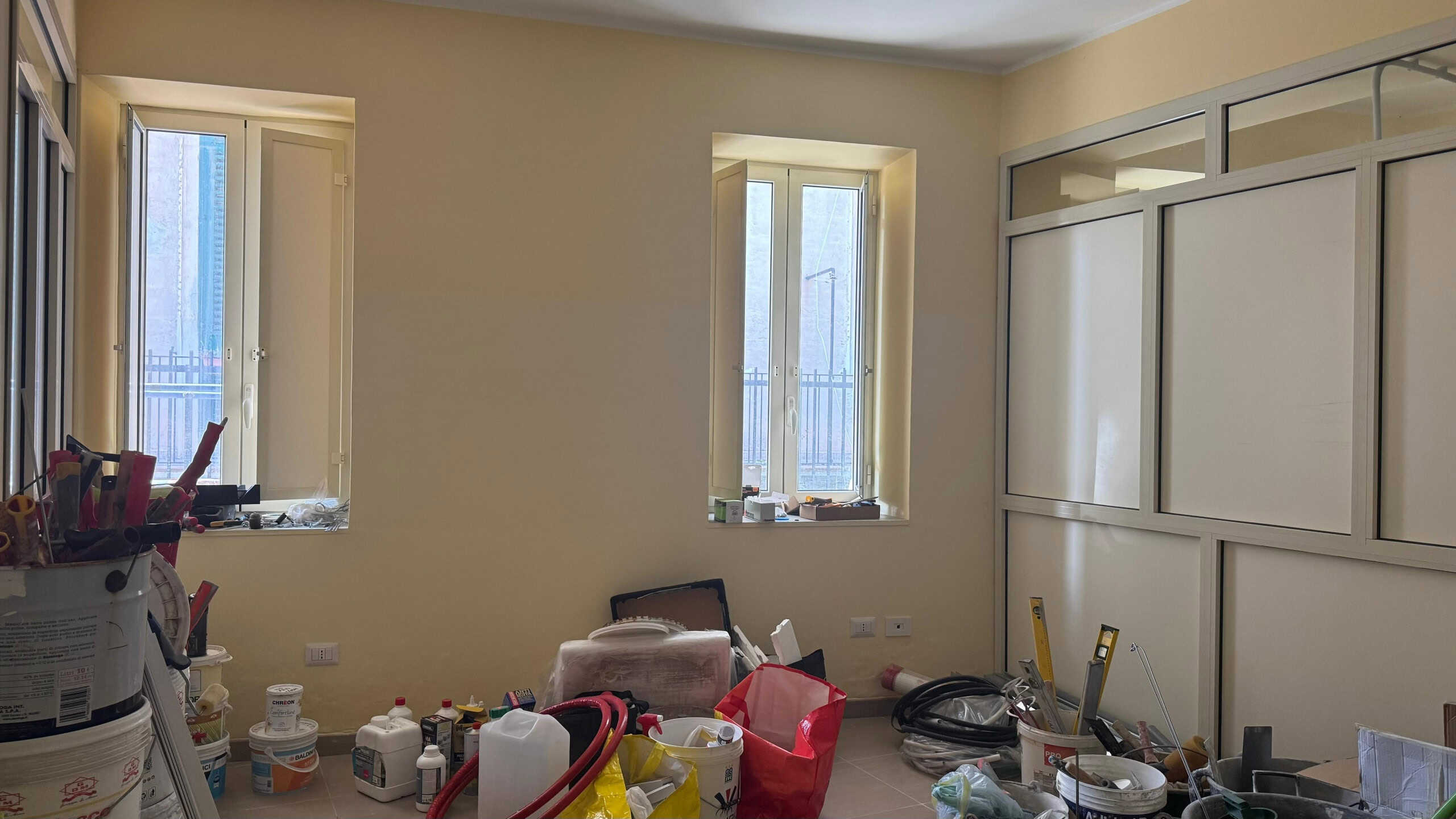 Cantiere in corso con lavori di ristrutturazione, attrezzi e materiali per il miglioramento degli interni, illuminato da luce naturale attraverso due finestre.