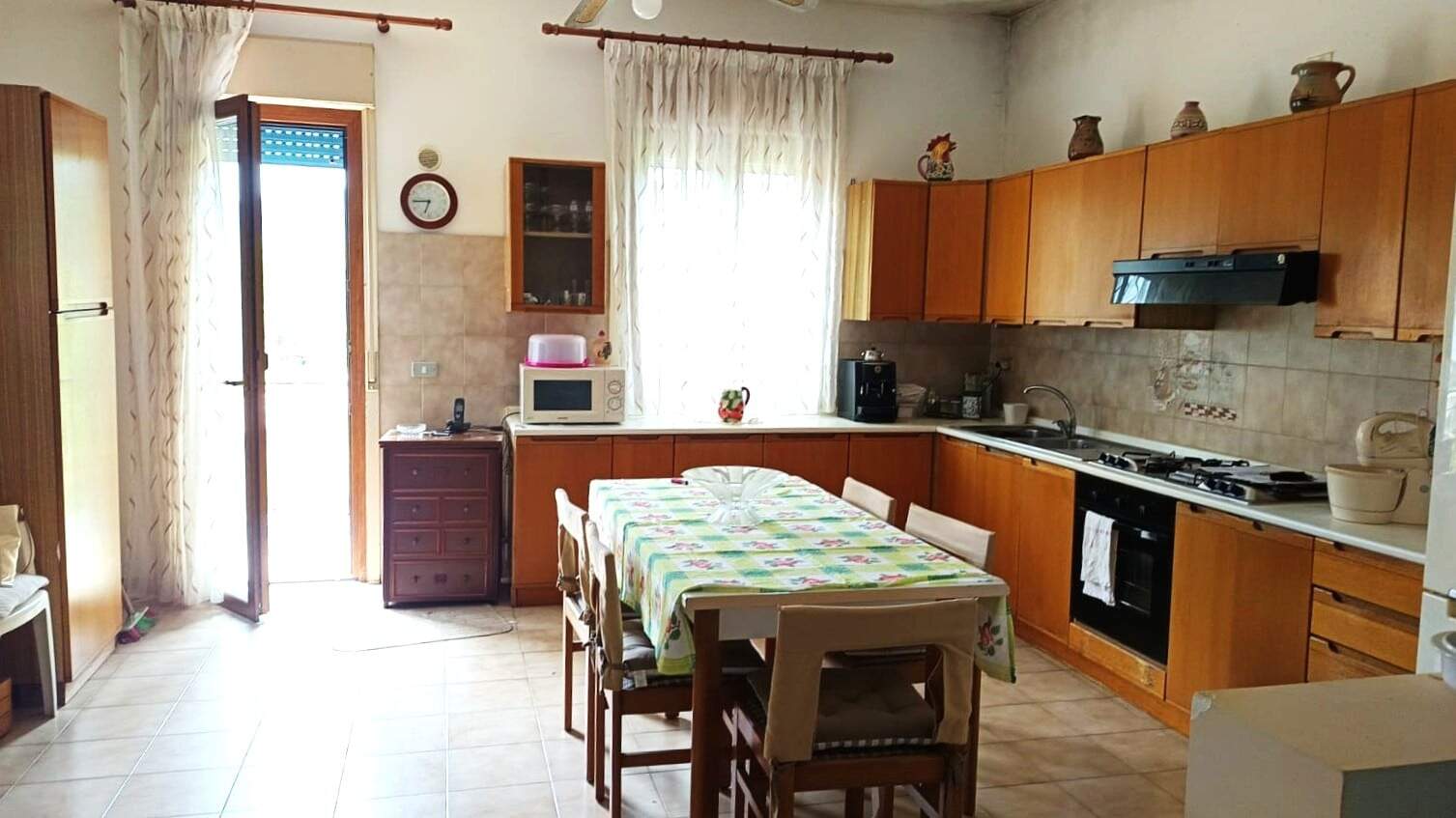 Spazio cucina abitabile in stile tradizionale con mobili in legno, tavolo da pranzo con tovaglia floreale e cucina attrezzata, luce naturale da finestra e porta finestra, ambiente accogliente e funzionale.