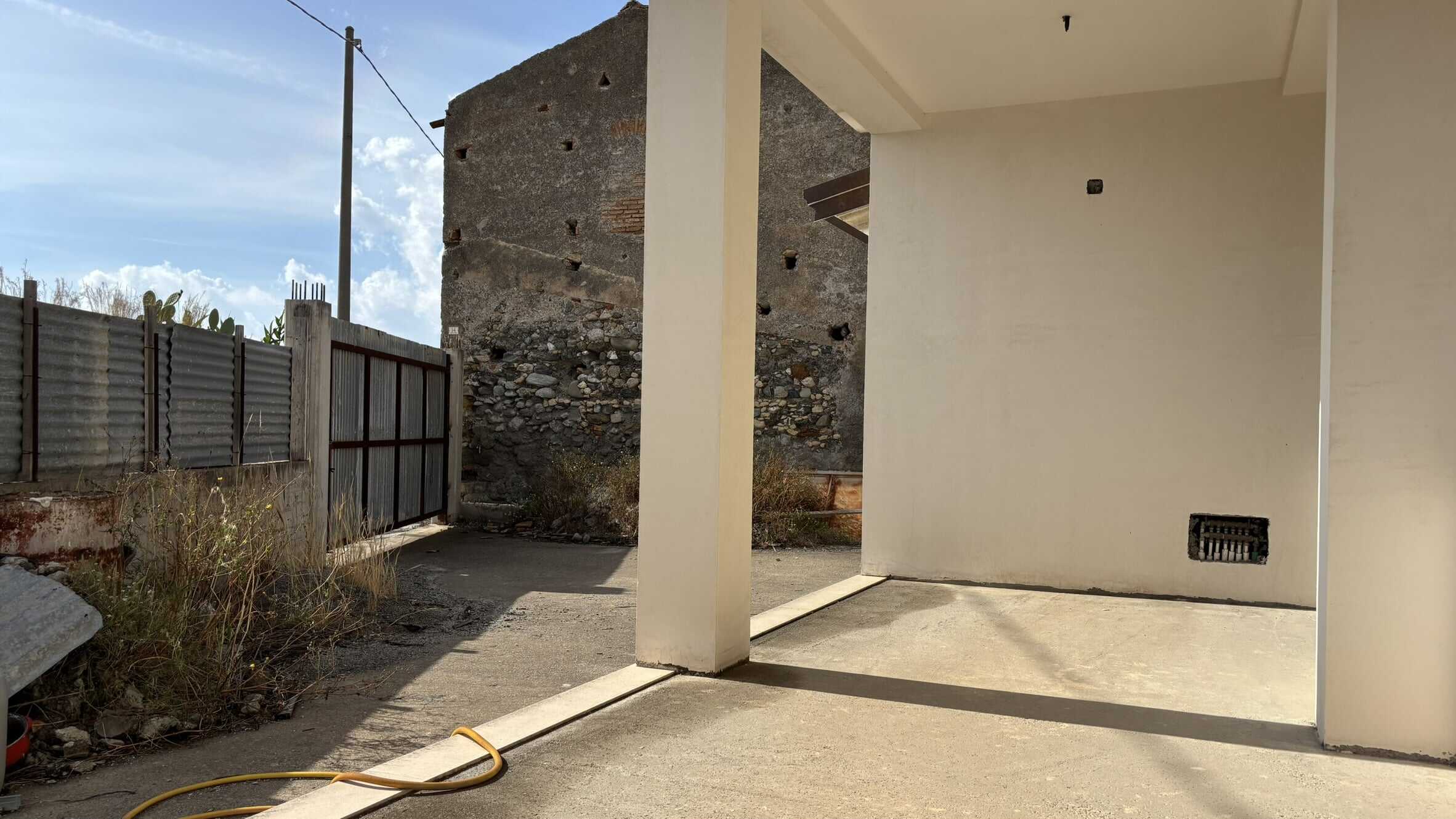Fondo: Finiture esterne di una casa moderna con spazio esterno e recinzione in metallo. La foto mostra l'area di un garage o portico in costruzione con pareti nuove e pavimento in cemento.