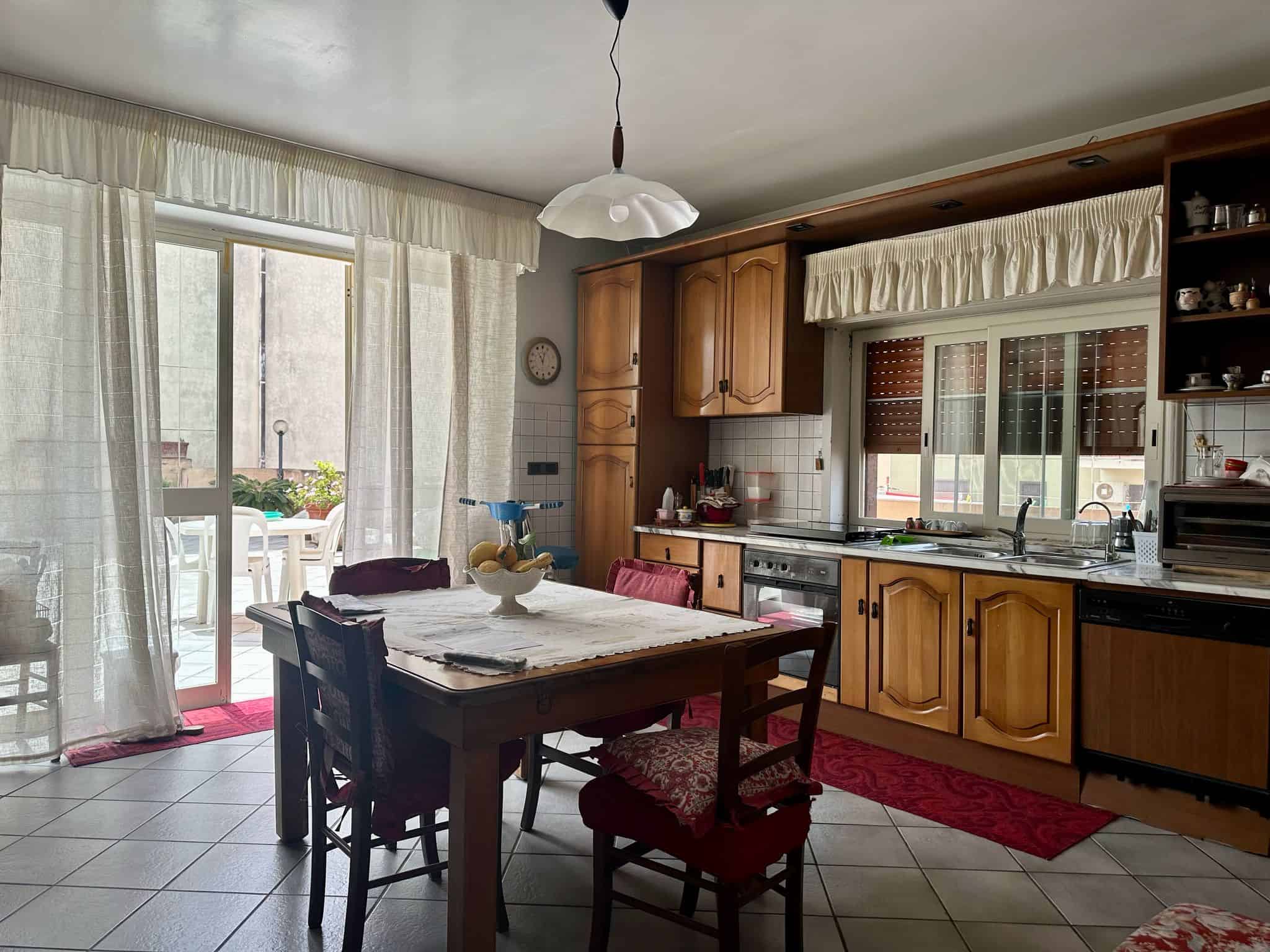 Vecchia cucina abitabile con mobili in legno, tavolo da pranzo, sedie e una vista sulla veranda esterna con balcone e sedie, ampie finestre con tende e illuminazione naturale.