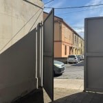 Porta di ingresso bifold all'aperto in zona urbana, prospettiva dalla strada con edifici e auto, stile moderno e funzionale, ambiente residenziale e commerciale, sicurezza e praticità per accesso all'abitazione, fotografia di alta qualità, luce naturale.