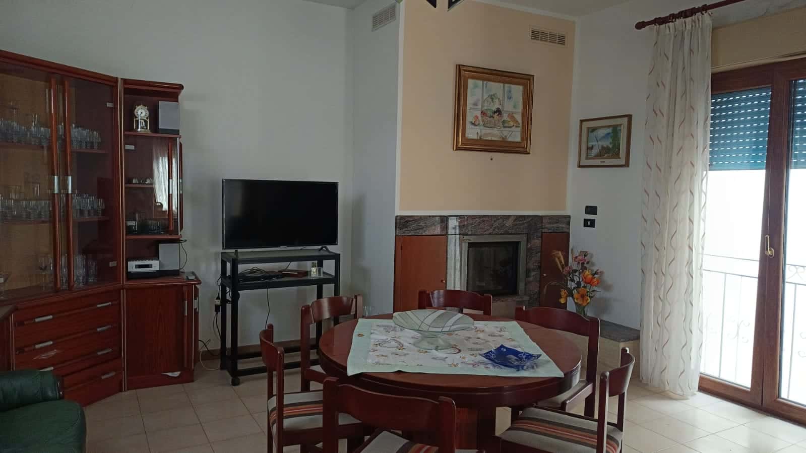 Mobili soggiorno con tavolo e divano vicino a camino e porta finestra con tende, arredamento tradizionale e ambiente luminoso, ideale per affitto o vendita immobiliare NextCasa.