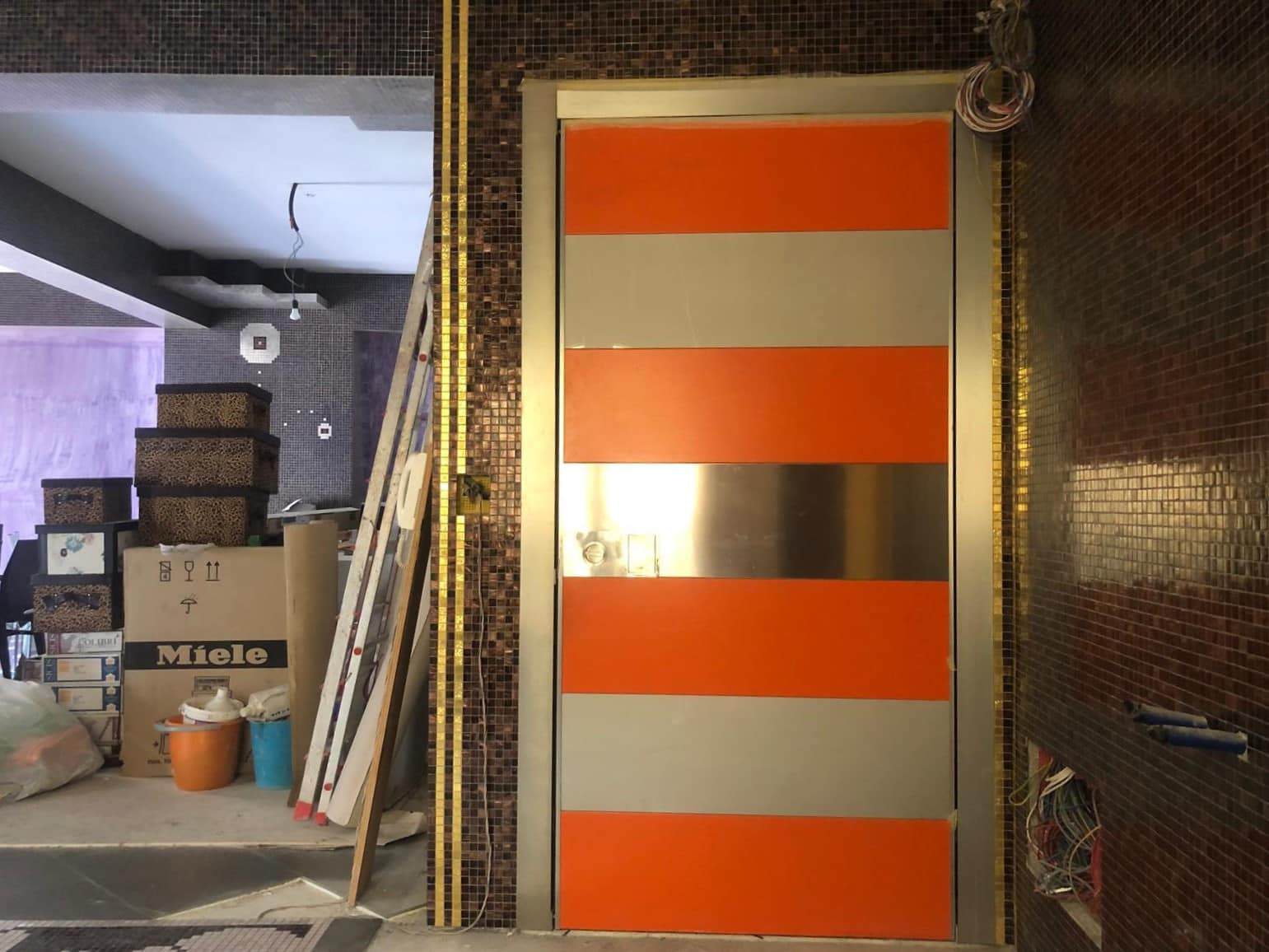 Porte da ascensore con profili in acciaio e pannelli in colore arancione e grigio, installazione in corso, ambiente commerciale o residenziale, dettagli di finitura in corso d'opera.