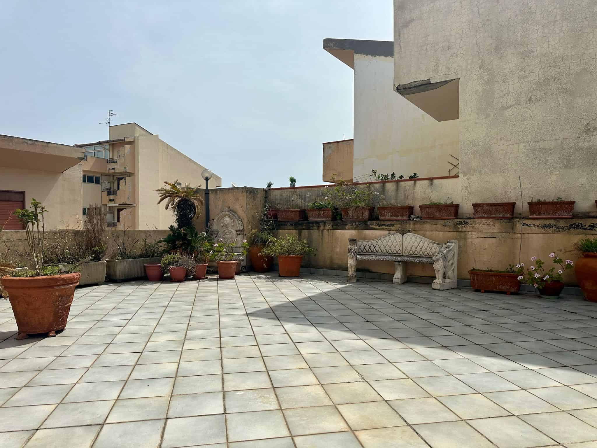 Terrazza balcone urbano con piante in vaso, panchina in pietra e vista su edifici in stile mediterraneo, ideale per momenti di relax all'aperto; ambiente residenziale luminoso e accogliente.