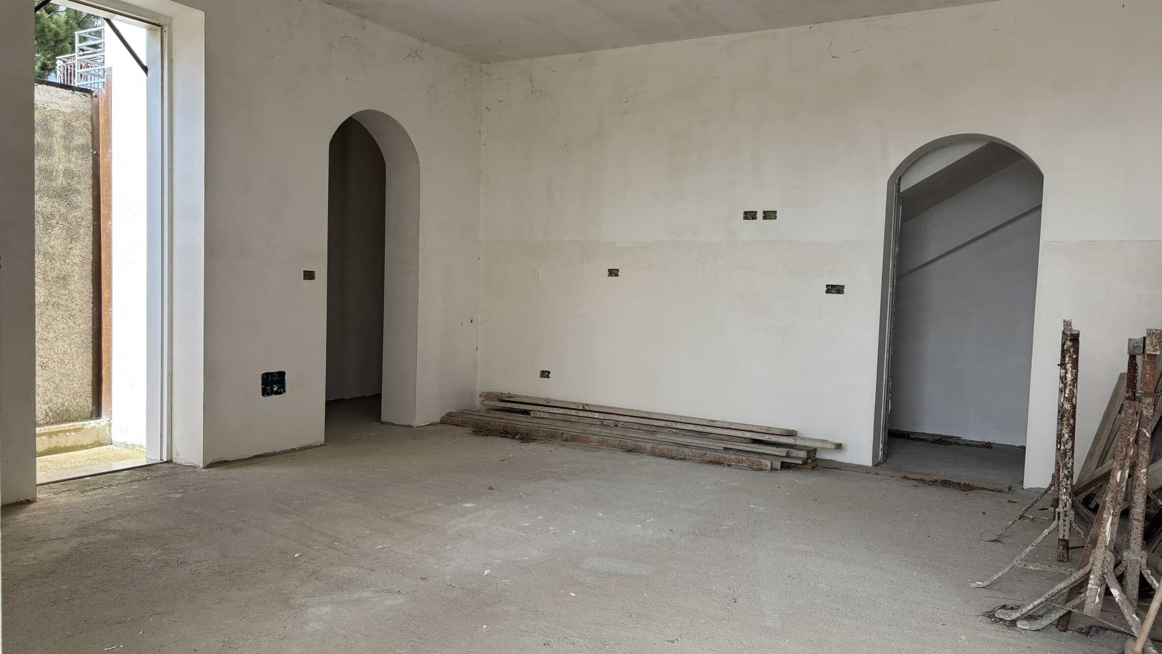 Stanza in fase di ristrutturazione con pareti neutre, porta ad arco e finestra grande per luce naturale, pronto per interventi di finitura e modernizzazione. Quando completata, sarà ideale per stanze residenziali accoglienti.