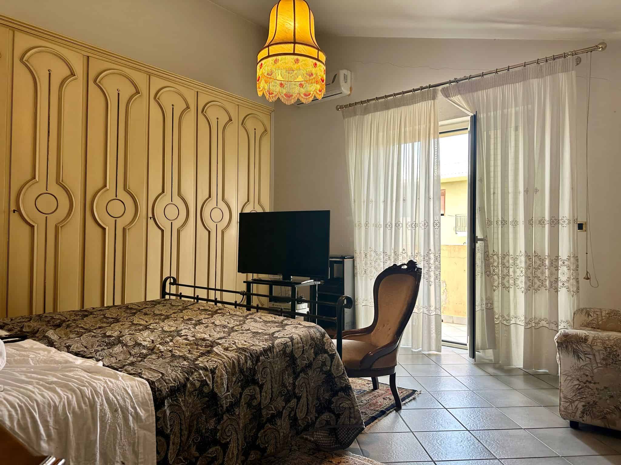 Stanza da letto spaziosa e luminosa con arredi classici, grande armadio a muro, divano e TV, luce naturale da porta finestra con tende leggere, ideale per case e appartamenti in vendita o affitto.