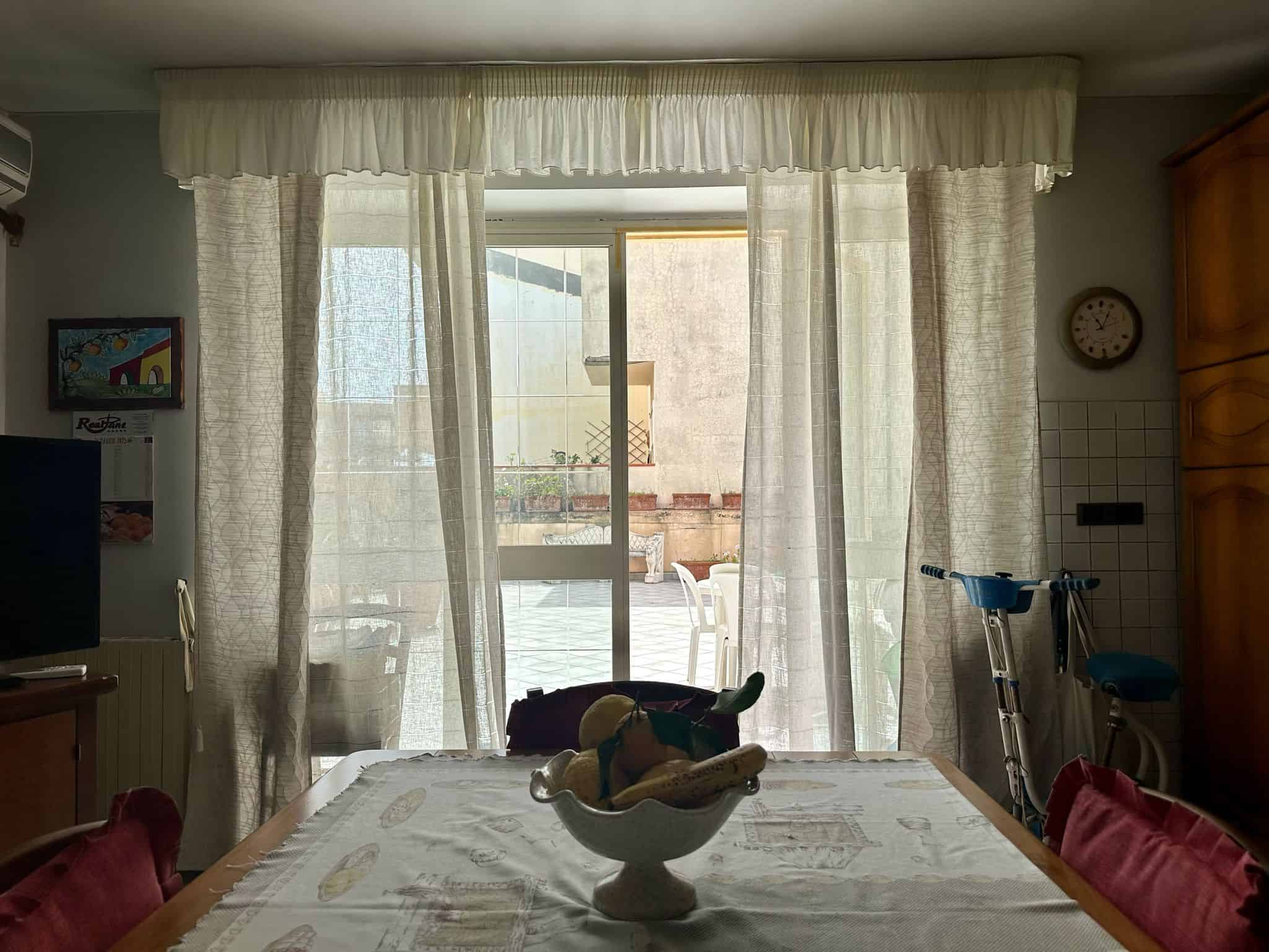 Finestra con tende trasparenti che apre su terrazza esterna, ambientazione domestica luminosa e accogliente, con mobili in legno, decorazioni e oggetti da cucina, ideale per ambienti di case e appartamenti.