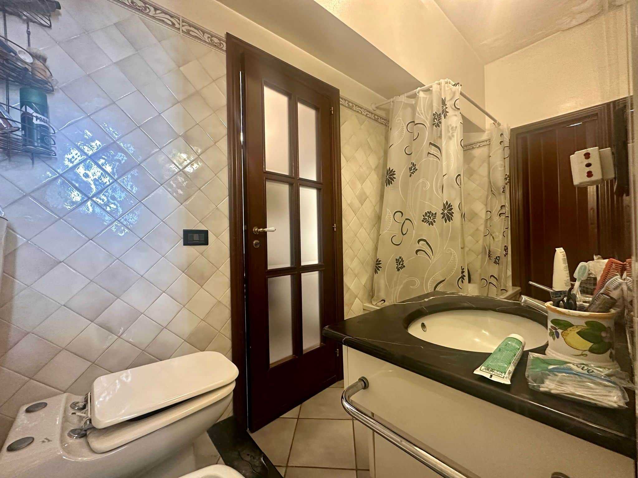 Bagno camino con piastrelle beige, lavabo con piano nero, doccia con tendina floreale, WC con sedile ribaltabile, acqua calda sanitaria, arredo pratico e funzionale, ideale per appartamento in vendita o affitto.