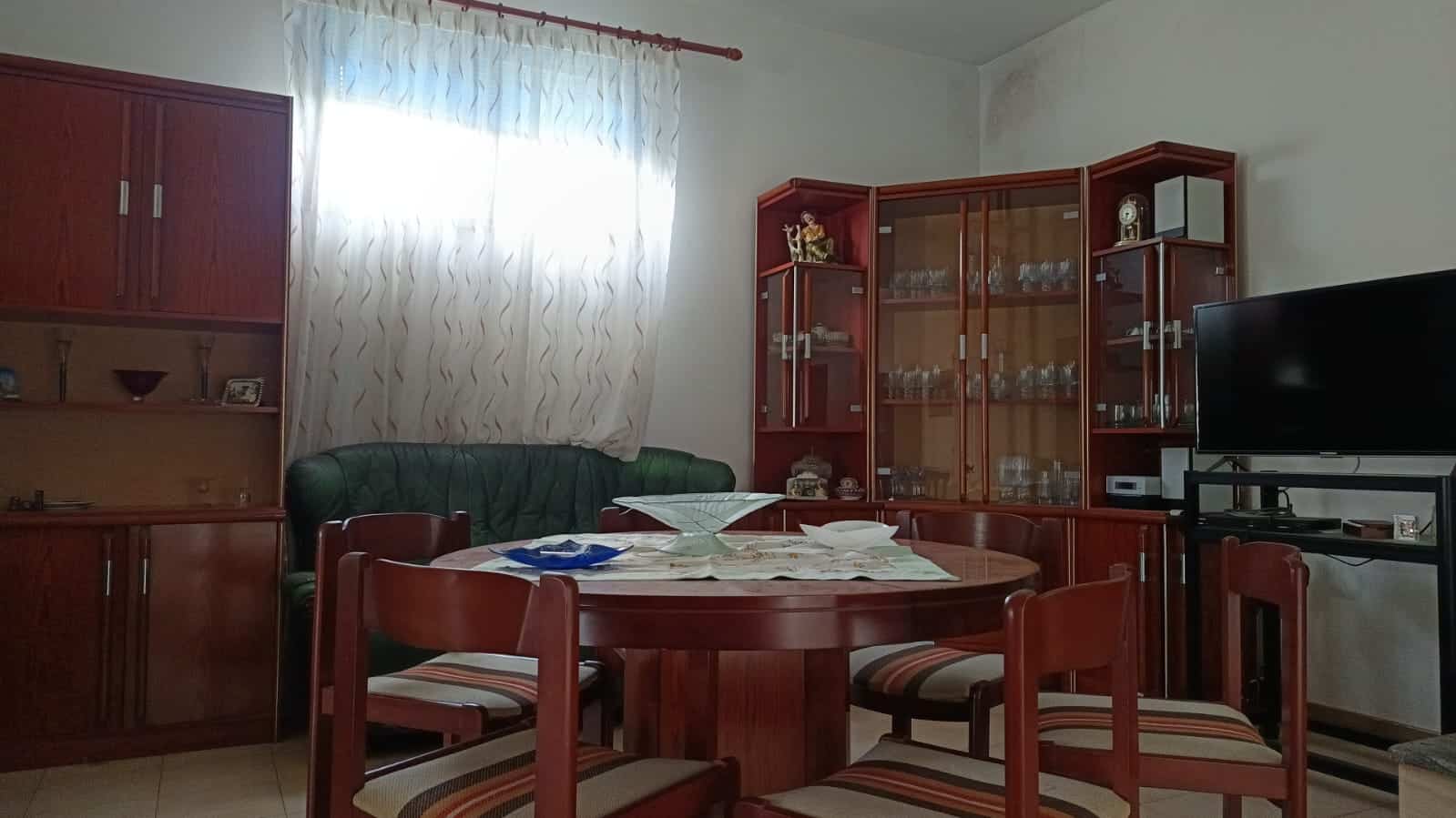 Arredamento soggiorno in legno, sala da pranzo con tavolo rotondo e sedie, credenze in legno con stoviglie e decorazioni, TV, atmosfera domestica e accogliente, esempio di design di interni abitazione.