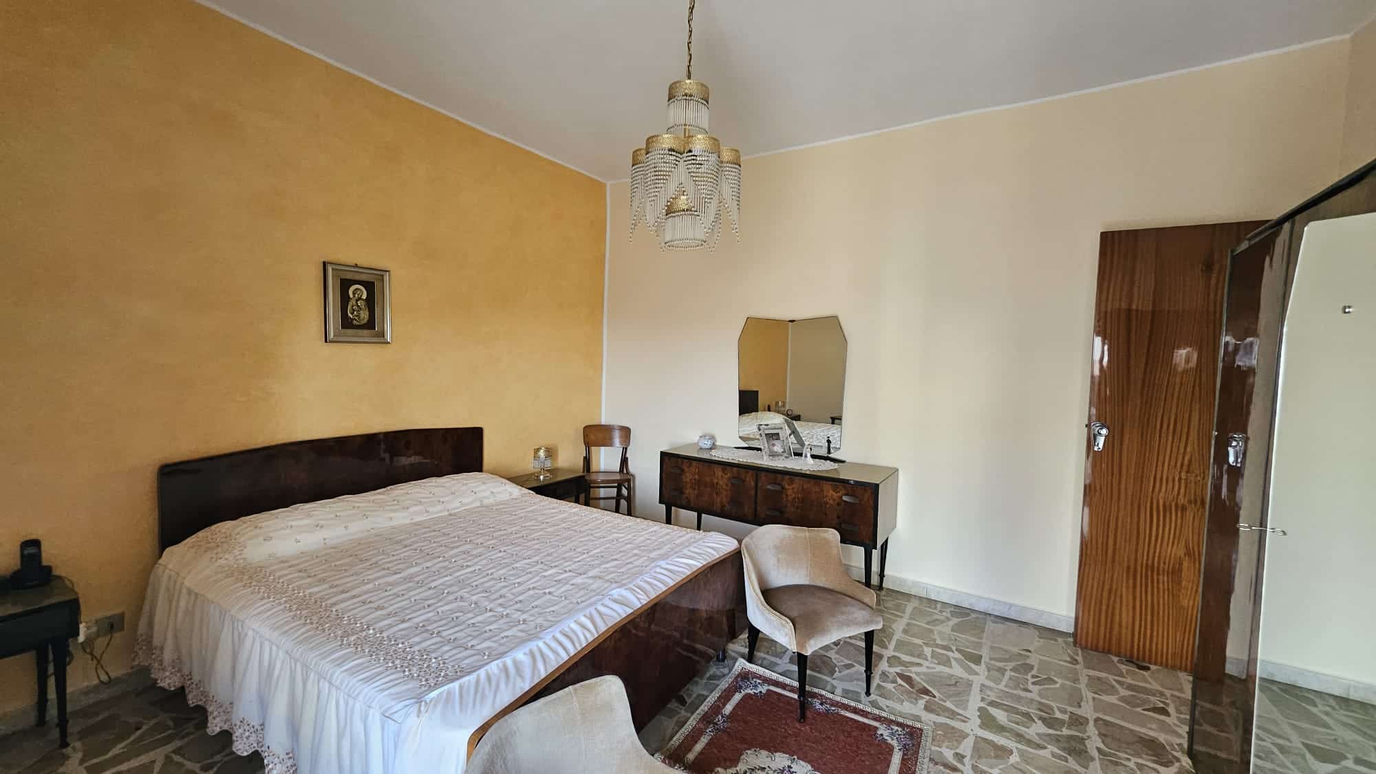 Letto matrimoniale in camera da letto con pareti gialle e arredi vintage, lampadario anni '70, specchio e comò in legno, spazio relax e decorazioni classiche, ideale per immobili di charme e appartamenti in vendita o affitto.